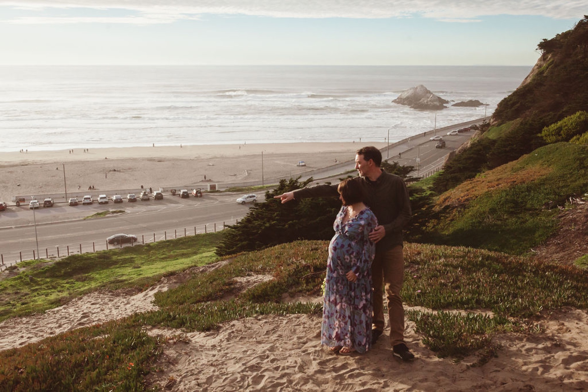 Expectant couple stands on a grassy hillside overlooking the beach, the man's hand gently cradling the woman's pregnant belly as golden afternoon light bathes the coastal landscape