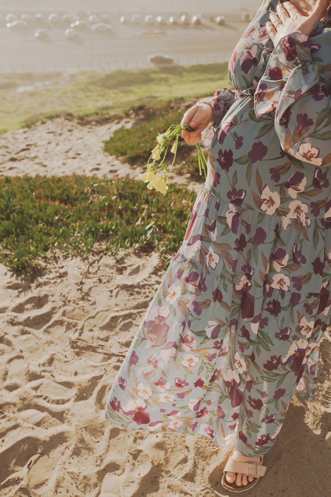 Pregnant woman in floral dress stands on sandy beach, cradling belly with both hands while gazing toward misty water and distant hills