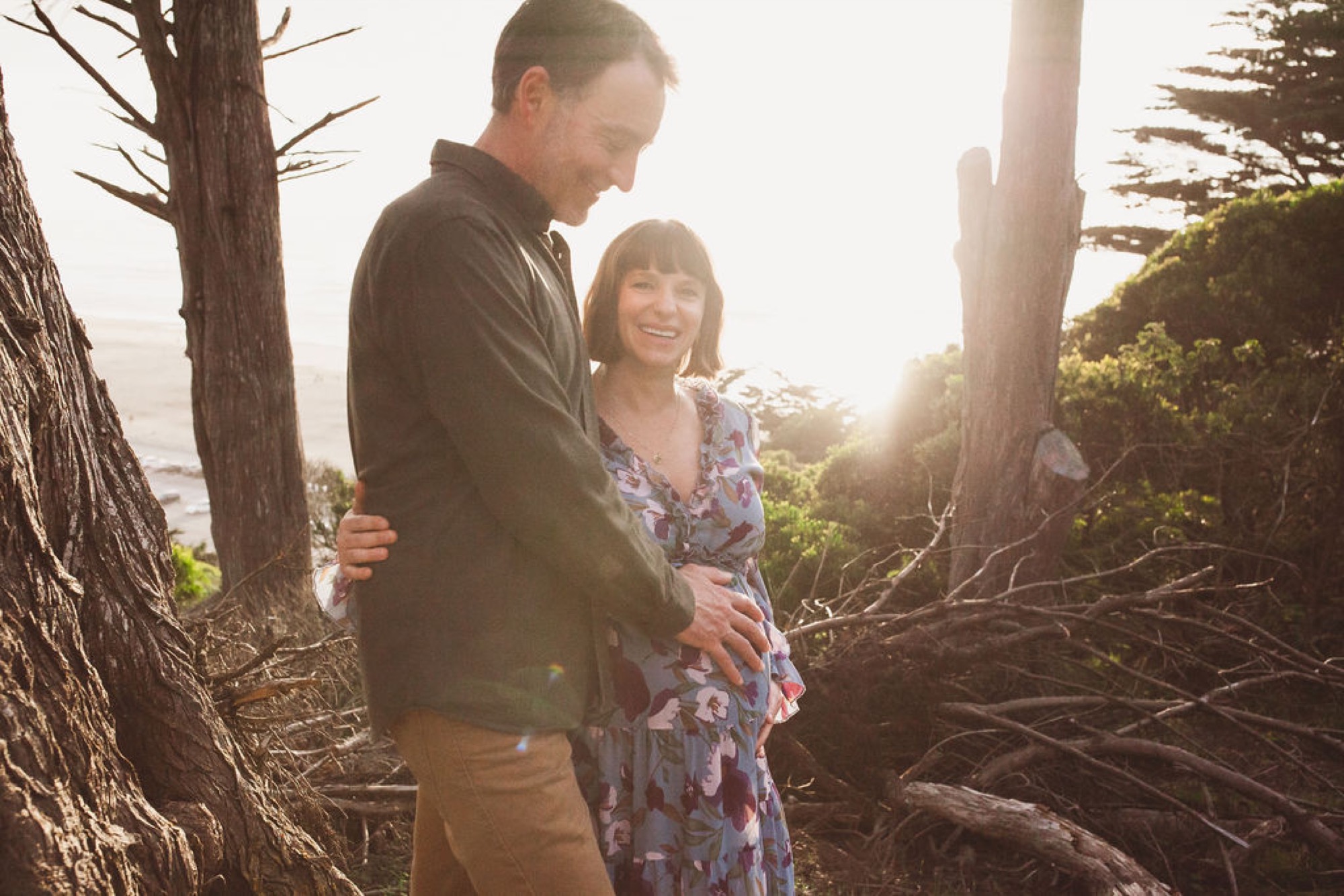 Expectant parents walk together through towering redwoods, golden afternoon light filtering through branches as the woman cradles her belly