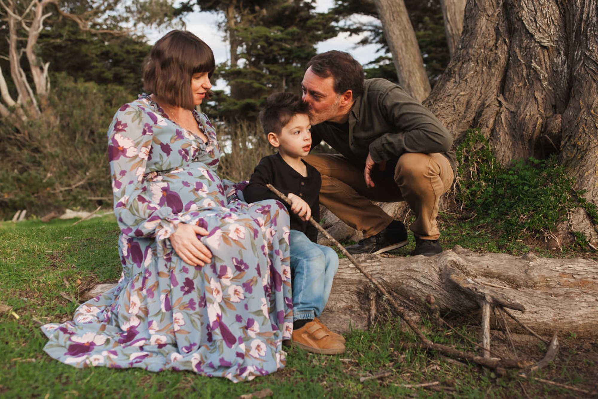 Pregnant woman in floral dress shares a tender moment with her partner and young son beside a weathered log beneath tall cypress trees