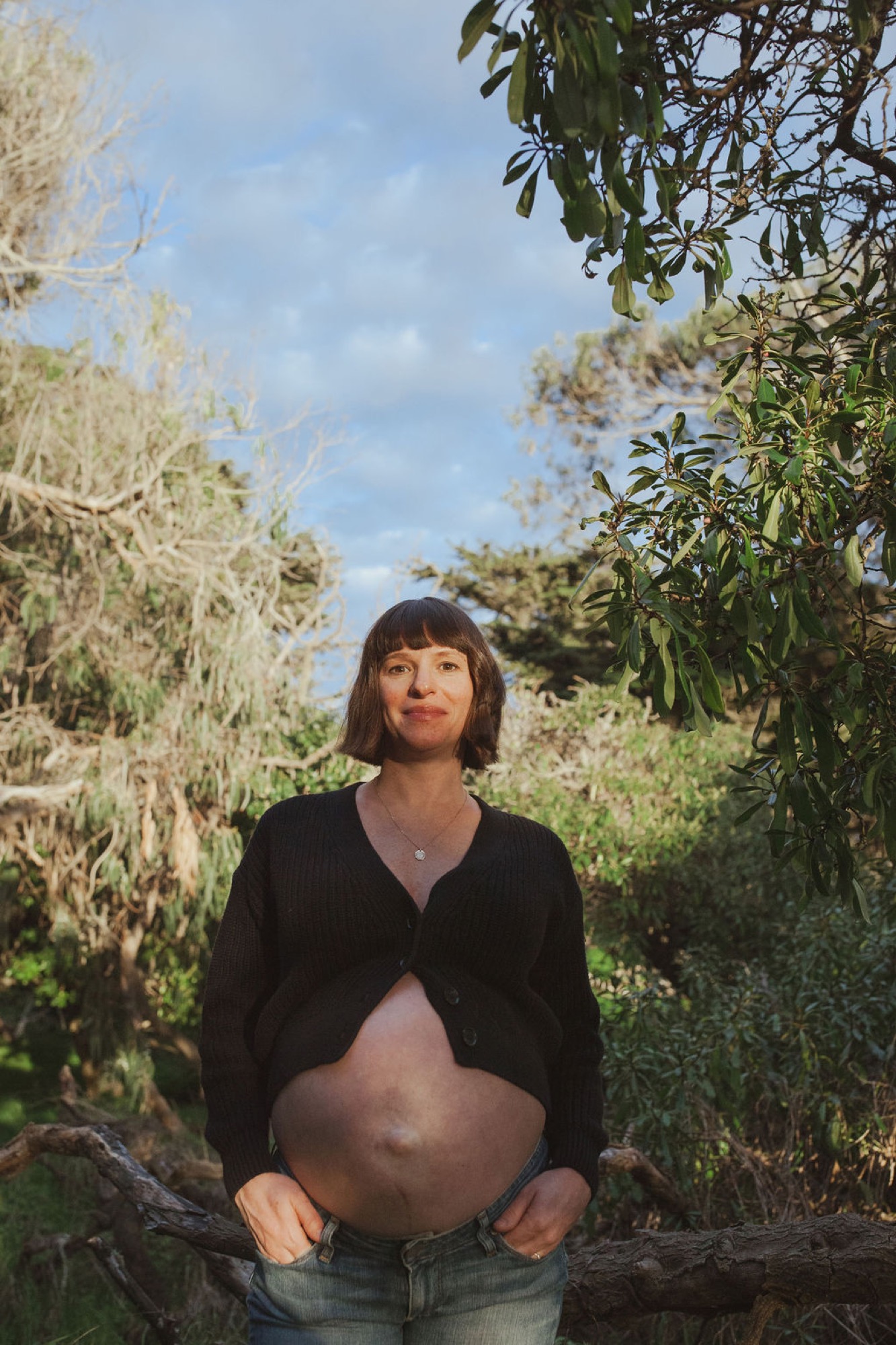 Pregnant woman in black cardigan stands among tall trees, gazing directly at camera with serene expression under dappled sunlight
