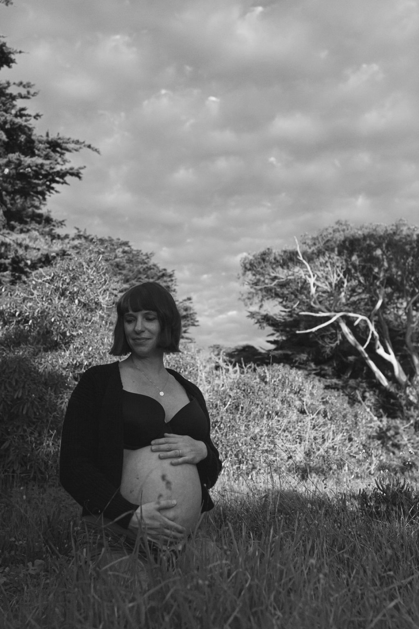 Pregnant woman in black cardigan stands contemplatively in scrubland, one hand cradling her belly, wind-twisted tree framing the overcast sky behind her