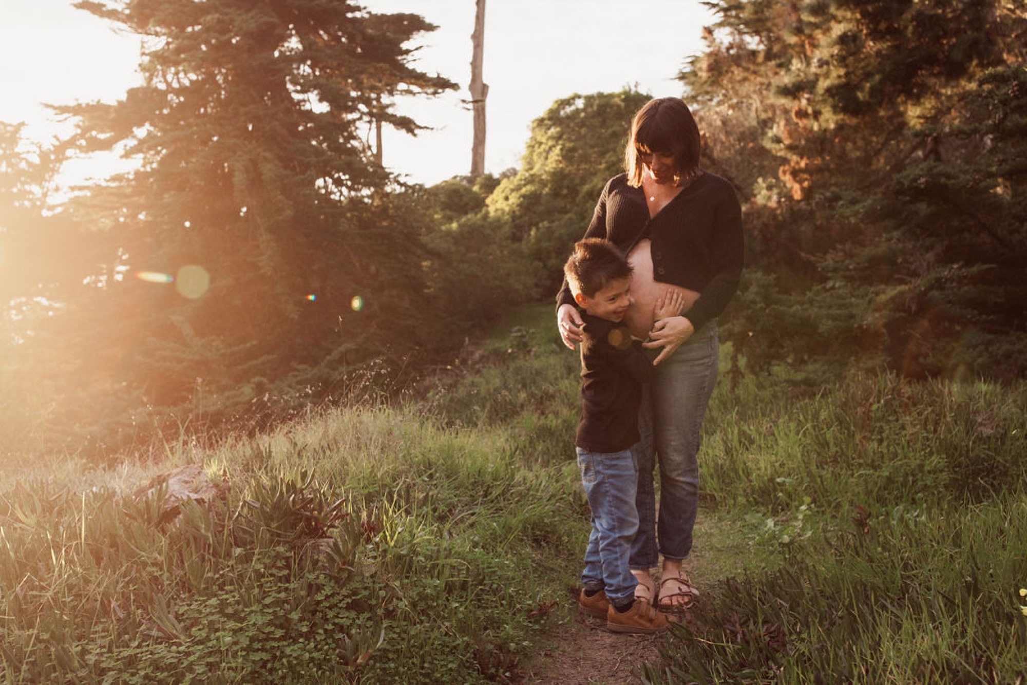 Woman holds infant close to her chest while standing on a sun-dappled forest path, golden light filtering through tall trees behind them