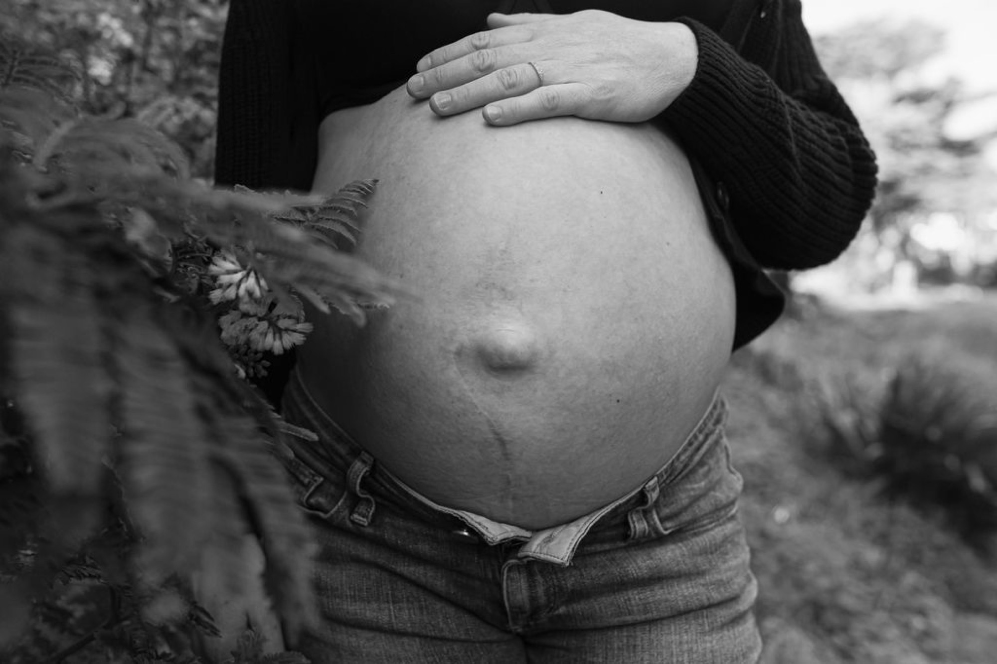 Pregnant woman cradling her bare belly with both hands in black and white, wearing denim and a dark sweater near coastal vegetation