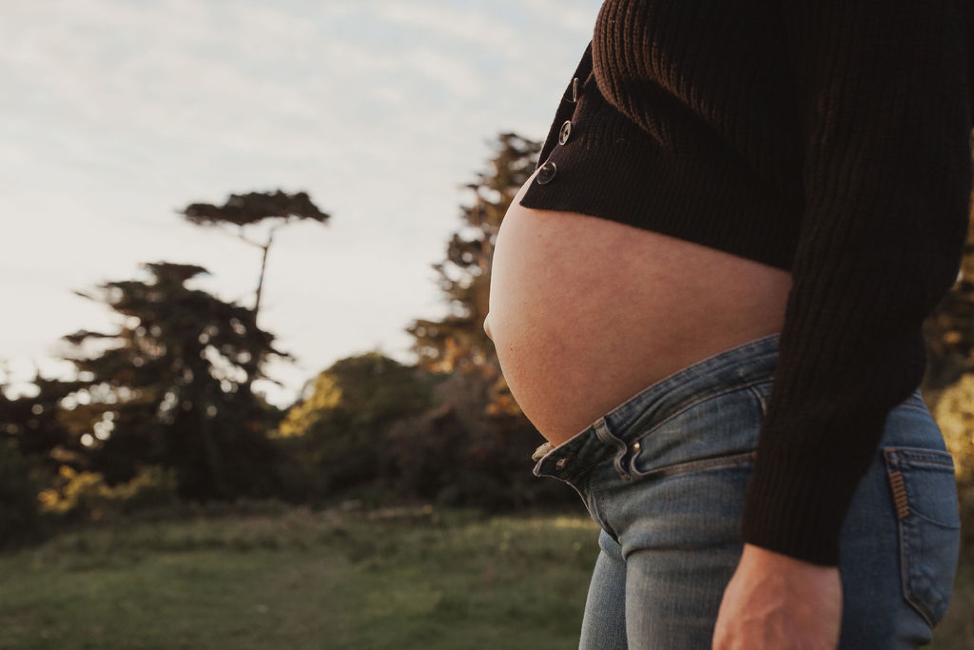 Pregnant woman in denim jeans profiles her bare belly against golden afternoon light, with windswept cypress trees silhouetted behind her