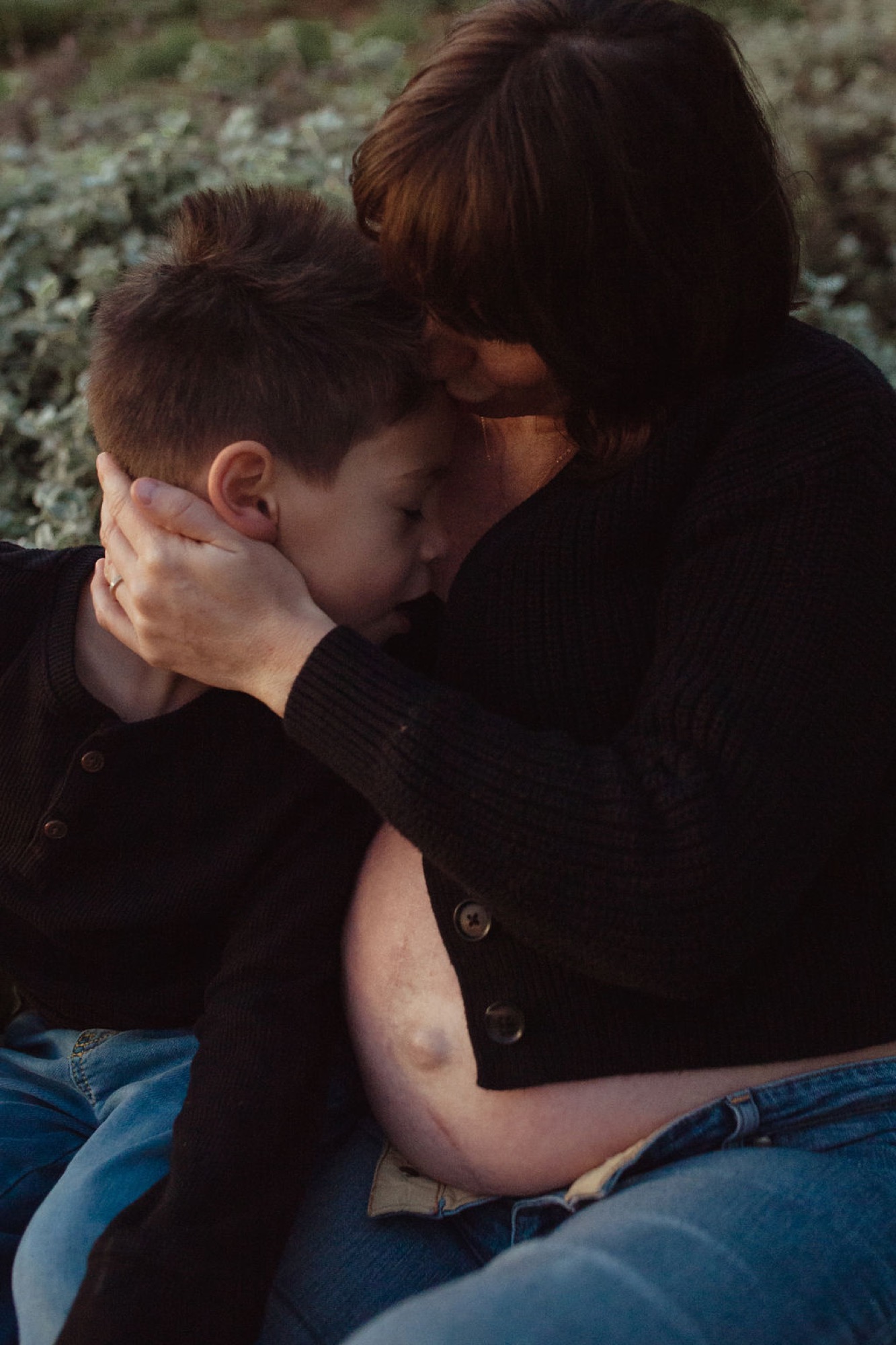 Two figures in dark clothing embrace intimately on ground with soft moss-covered rocks blurred in warm afternoon light