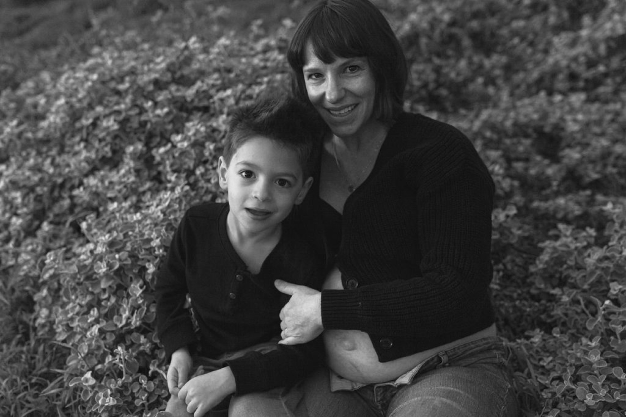 A woman and young boy sit together against lichen-covered rocks, both smiling warmly at the camera in soft natural light