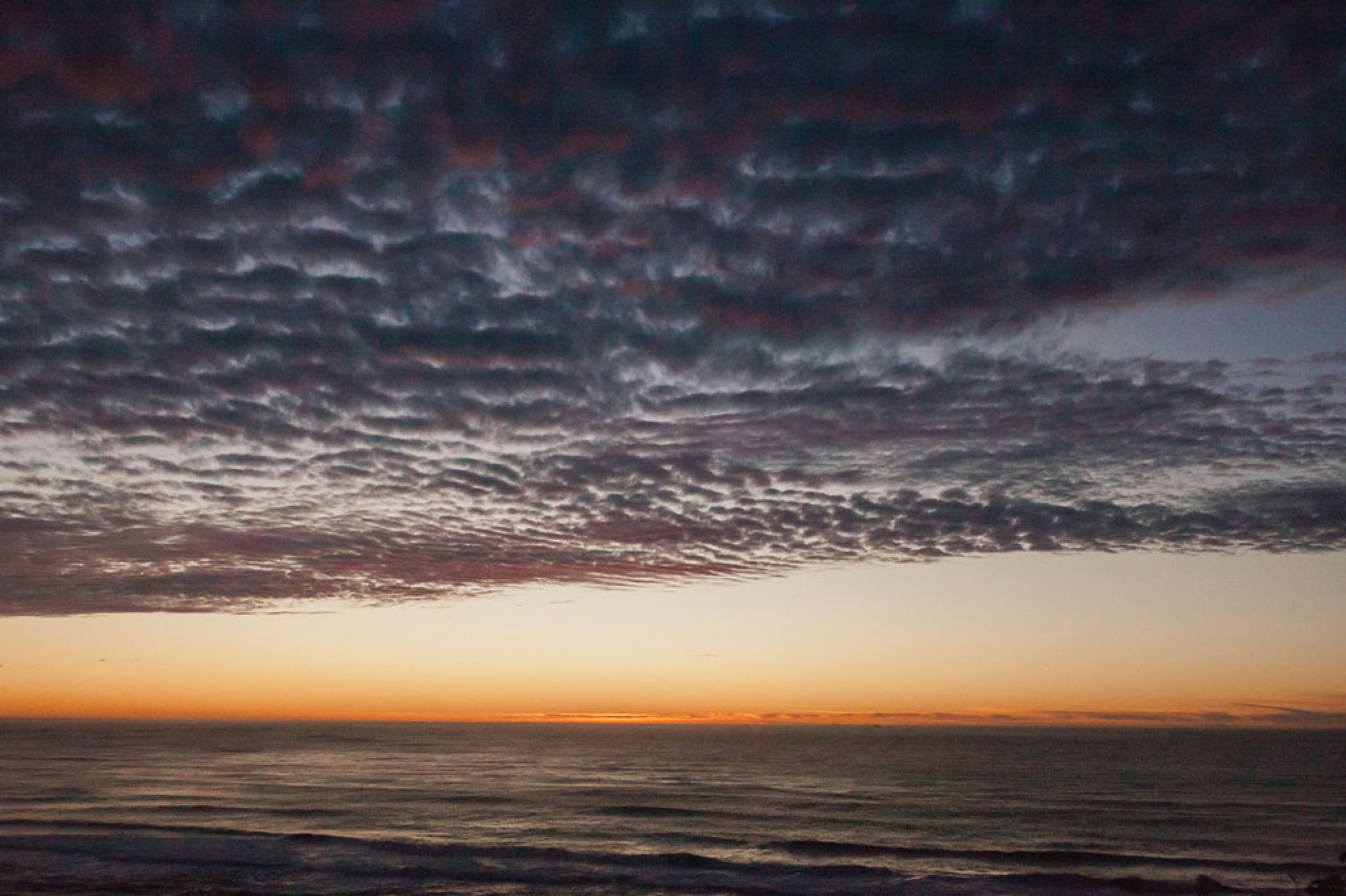 Dramatic textured clouds glow orange and pink above a dark ocean horizon at golden hour