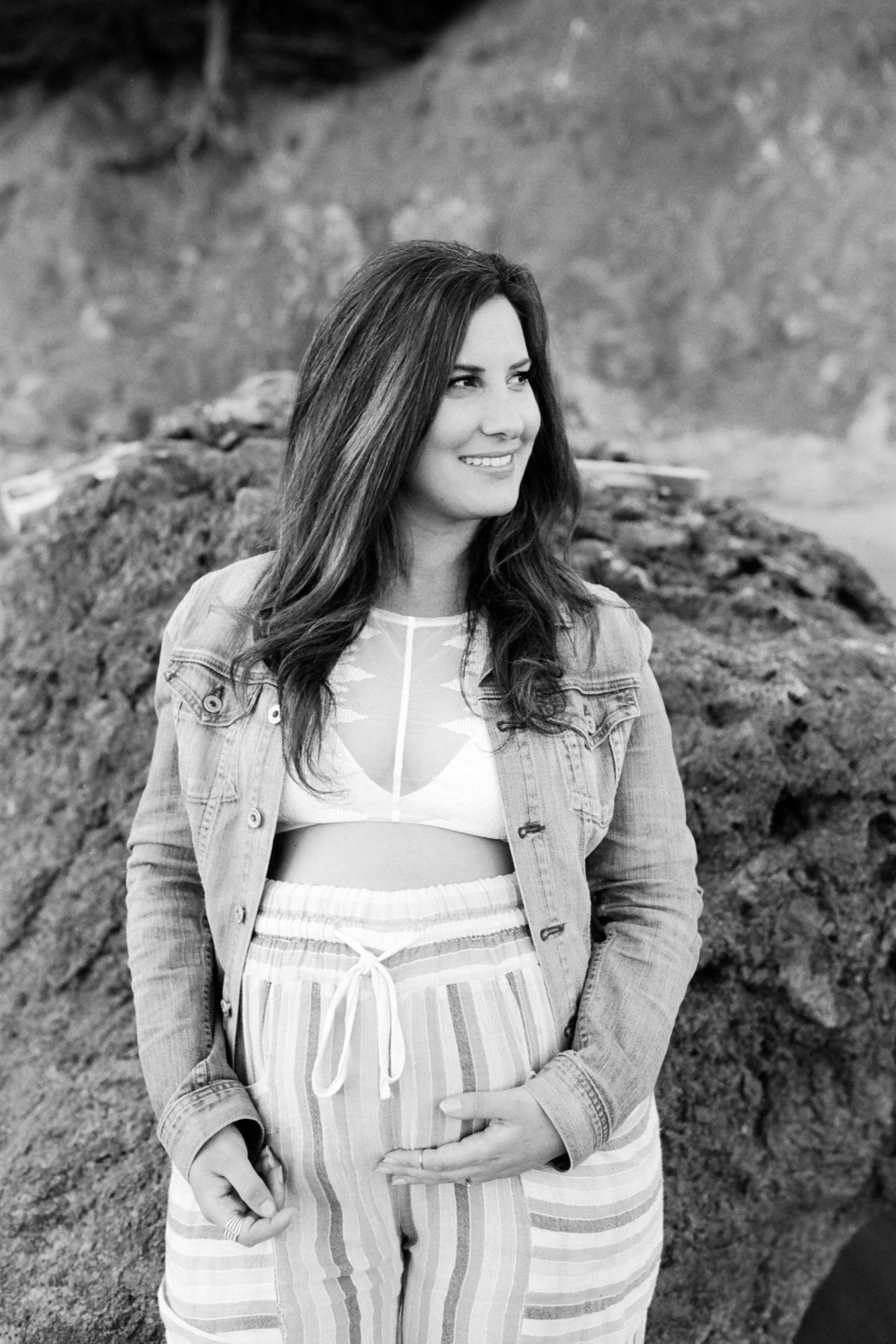 Expectant mother smiles warmly toward the horizon, one hand cradling her belly, rocky Muir Beach shore behind her