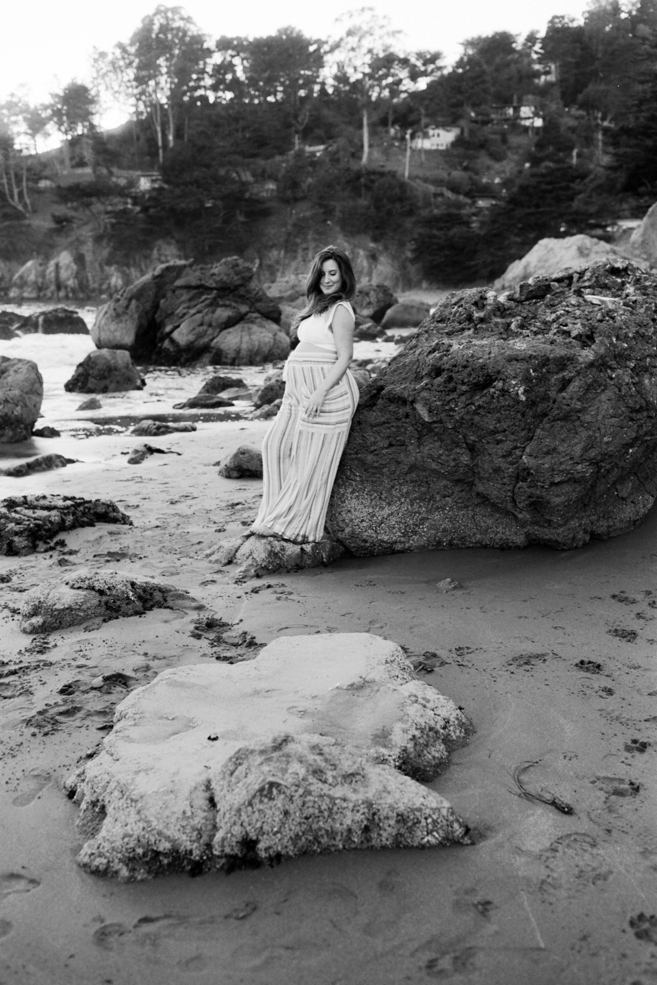 Pregnant woman in flowing white dress gazes downward beside massive boulders on the Muir Beach shoreline