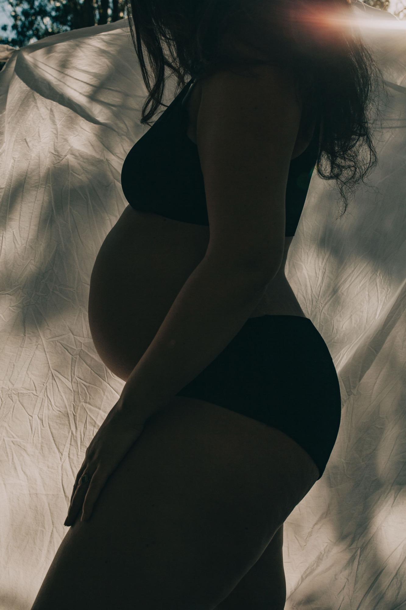 Pregnant woman silhouetted against sheer white fabric, backlit golden sunlight creating ethereal glow across her profile and rounded belly