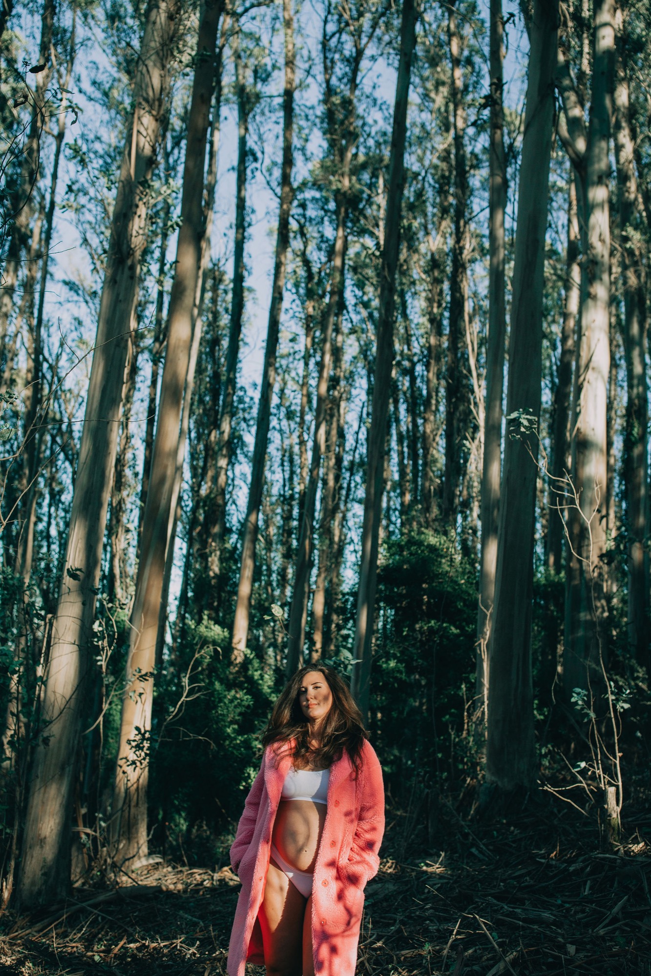 Pregnant woman in coral cardigan stands centered among towering eucalyptus trees, sunlight streaming through canopy onto her bare belly
