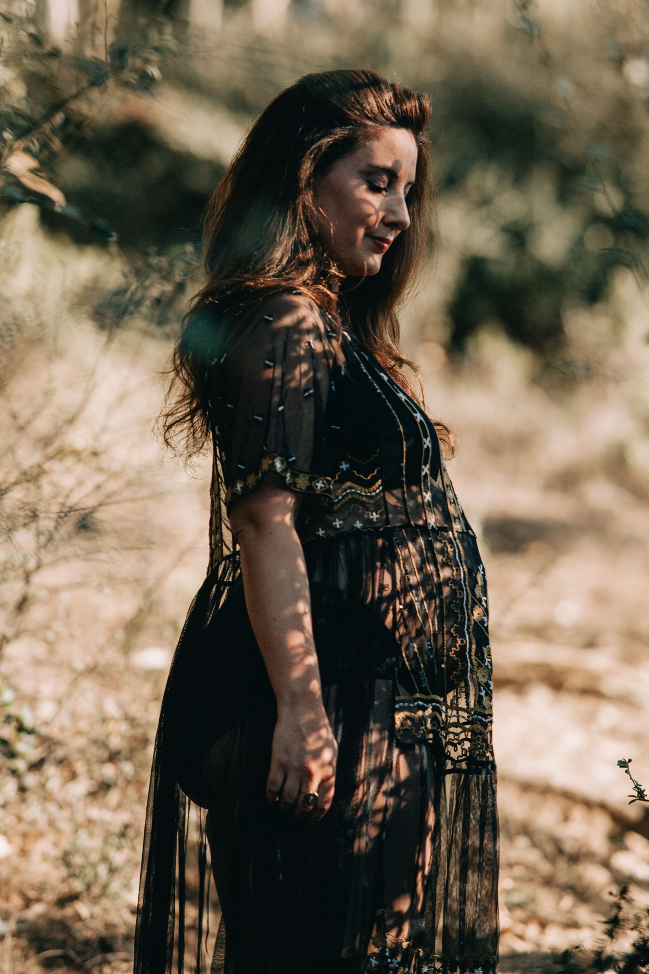 Pregnant woman in black embroidered dress gazes downward, dappled tree shadow patterning her silhouette against weathered stone backdrop