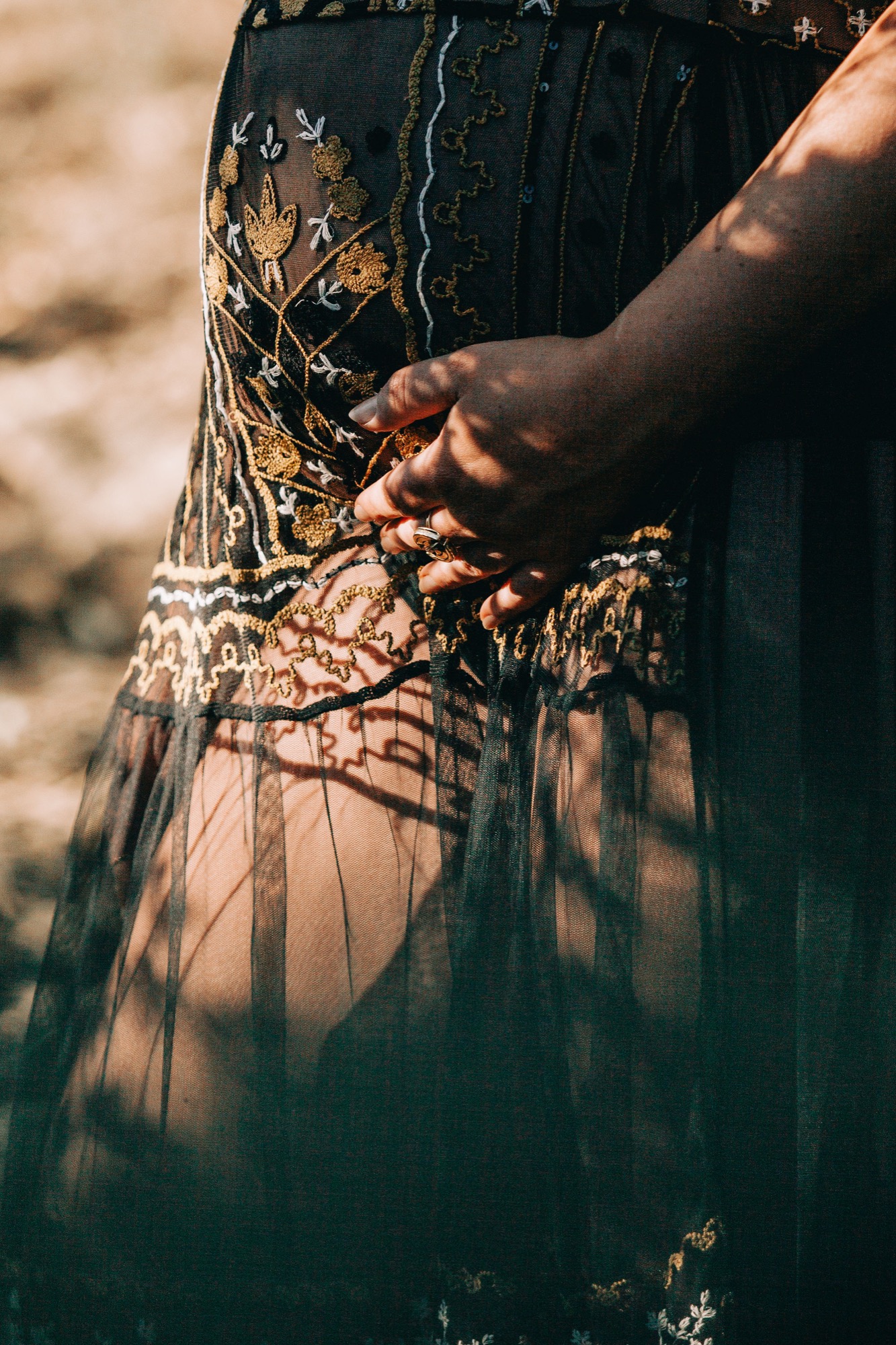 Pregnant woman in ornate black dress with gold embroidery cradling her belly, hands gently framing the bump as dappled sunlight creates shadow patterns across sheer fabric