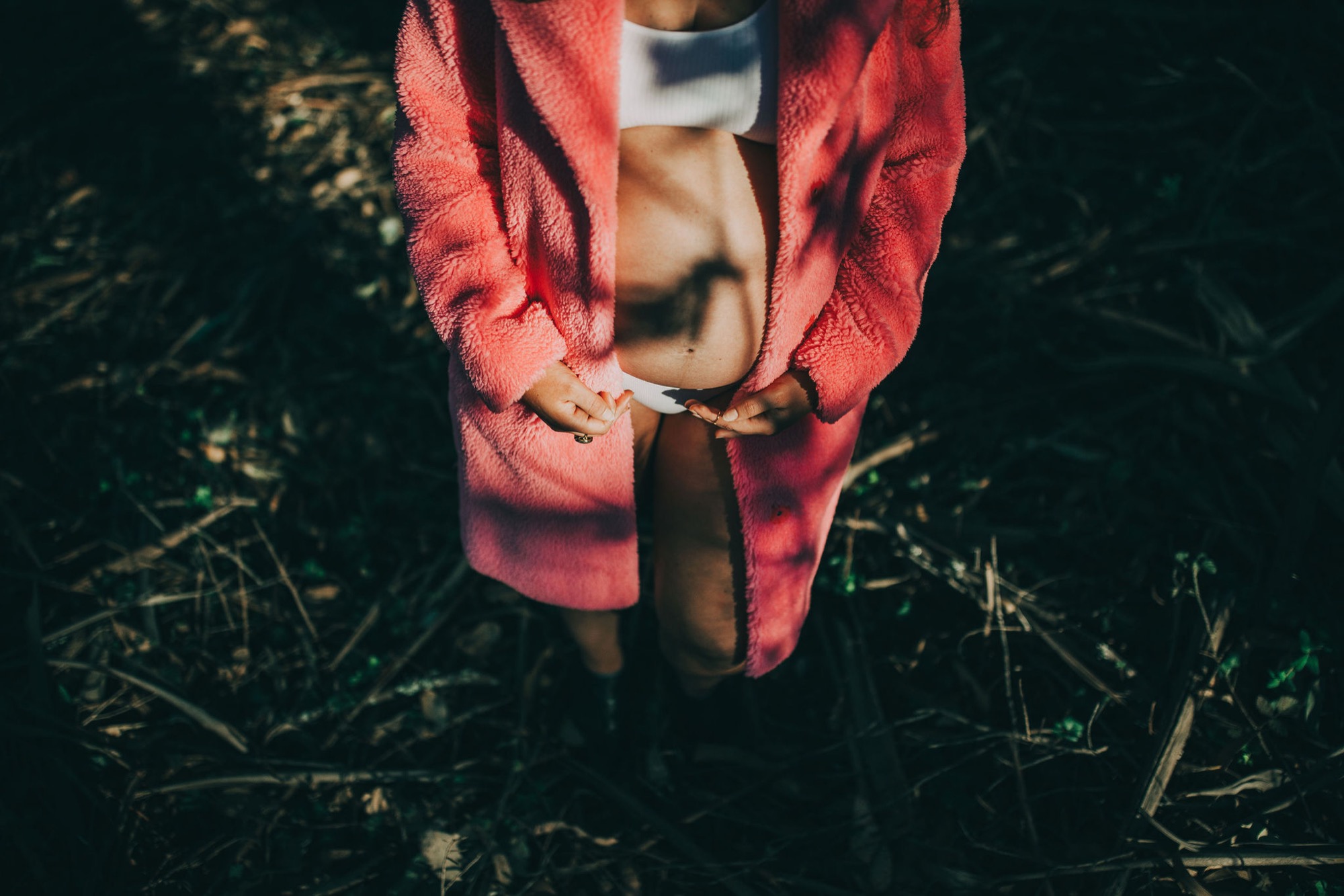 A pregnant woman in a vibrant coral wrap cradles her belly while standing on dark earth and dried grass, her white shirt peeking through