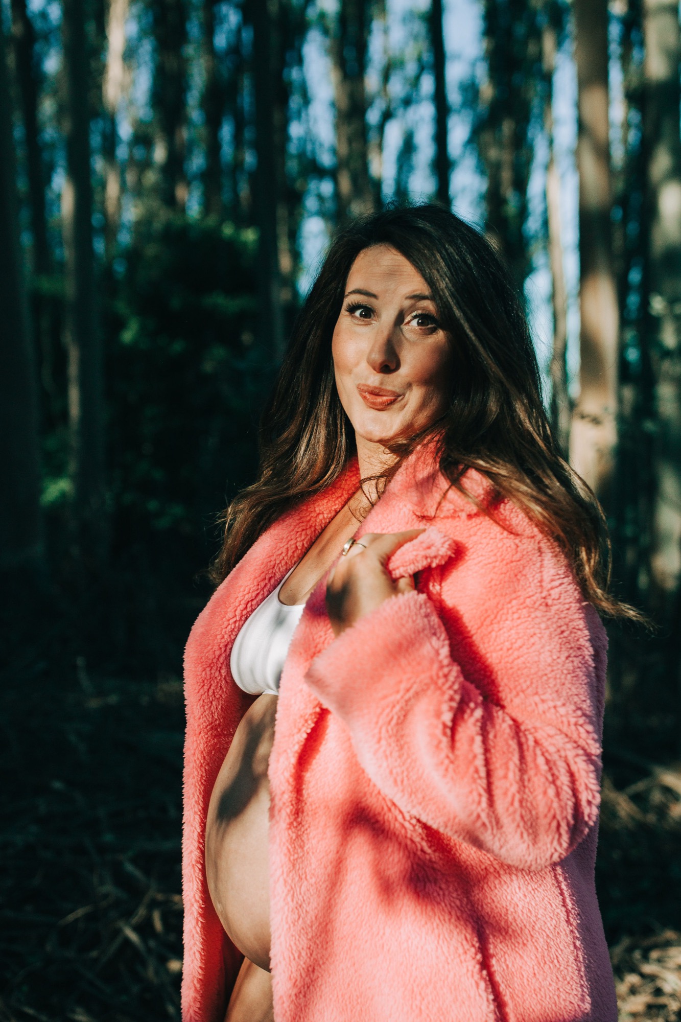 Pregnant woman in coral knit cardigan gazes over her shoulder among towering birch trees, warm light falling across her face