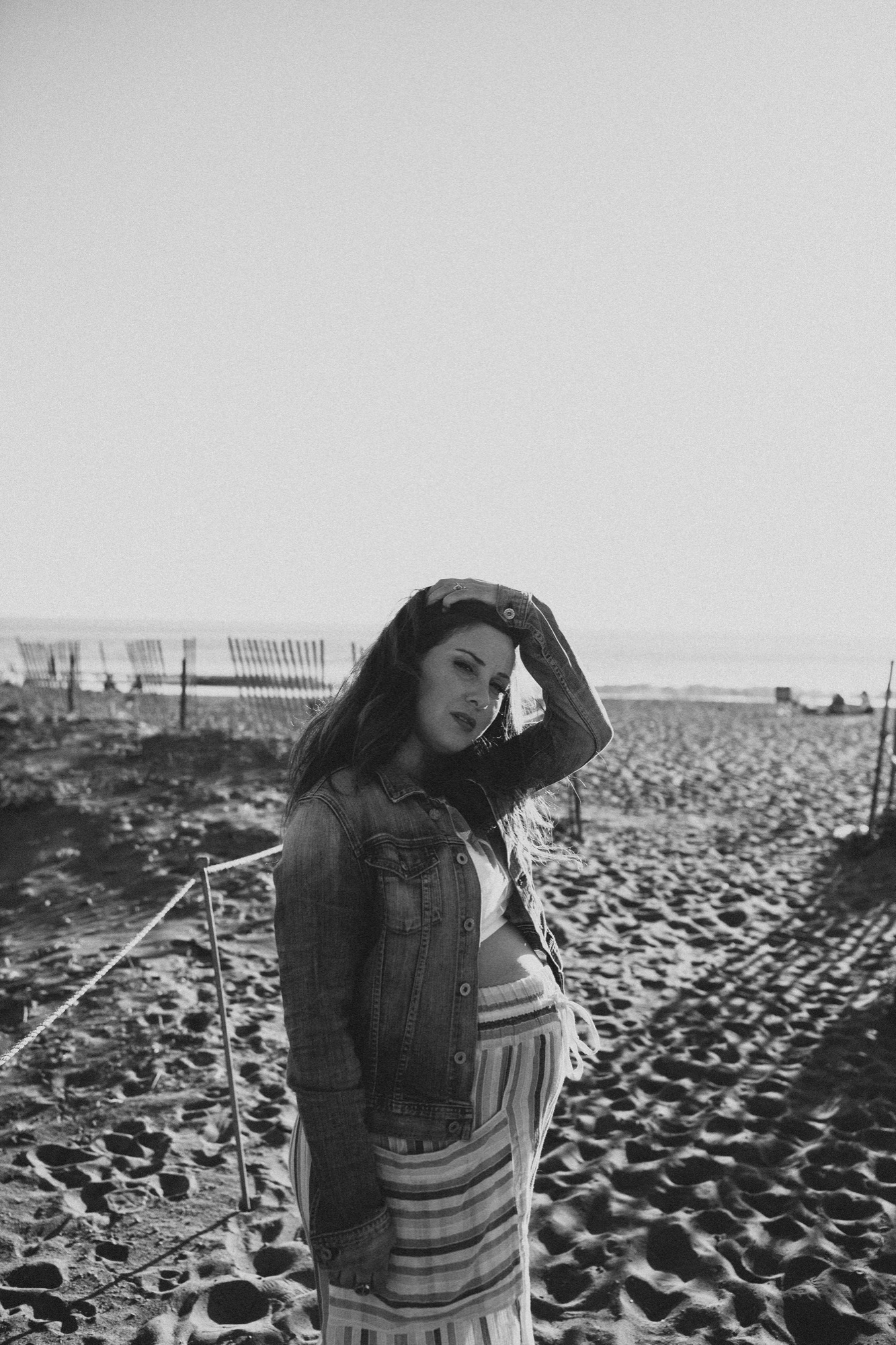 Pregnant woman in denim jacket gazes downward while cradling her belly in a vast plowed field under overcast sky