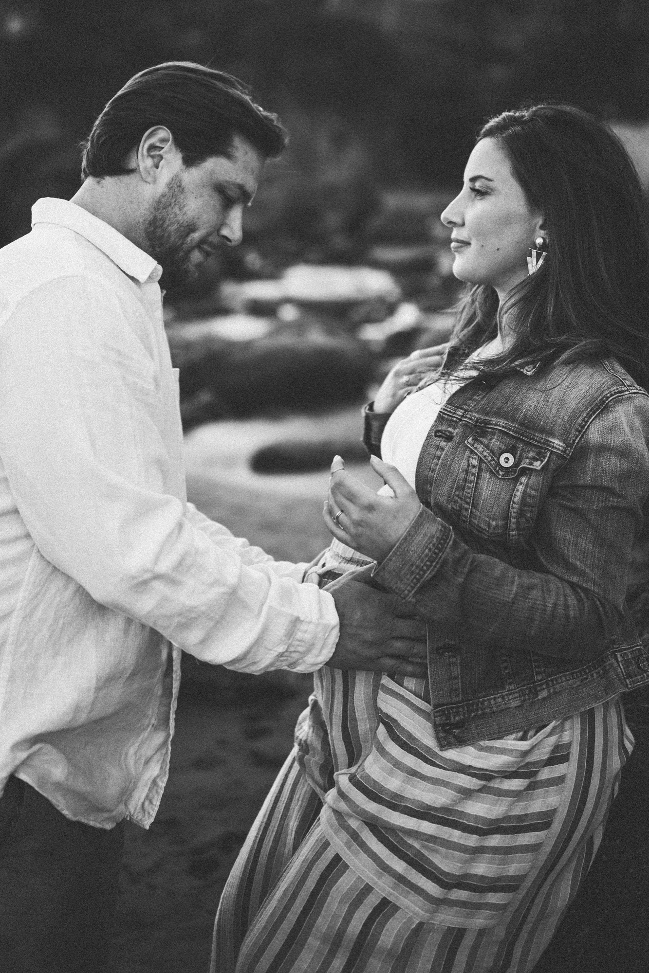 A bearded man in a white shirt gazes down at his pregnant partner's belly while she looks toward the camera, their hands connected in an intimate moment against a blurred natural backdrop