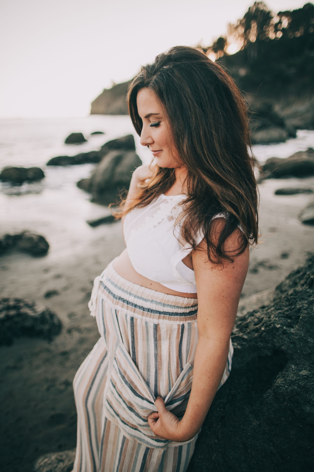 Pregnant woman in striped pants gazes downward at the rocky beach at golden hour, hand resting gently on her belly