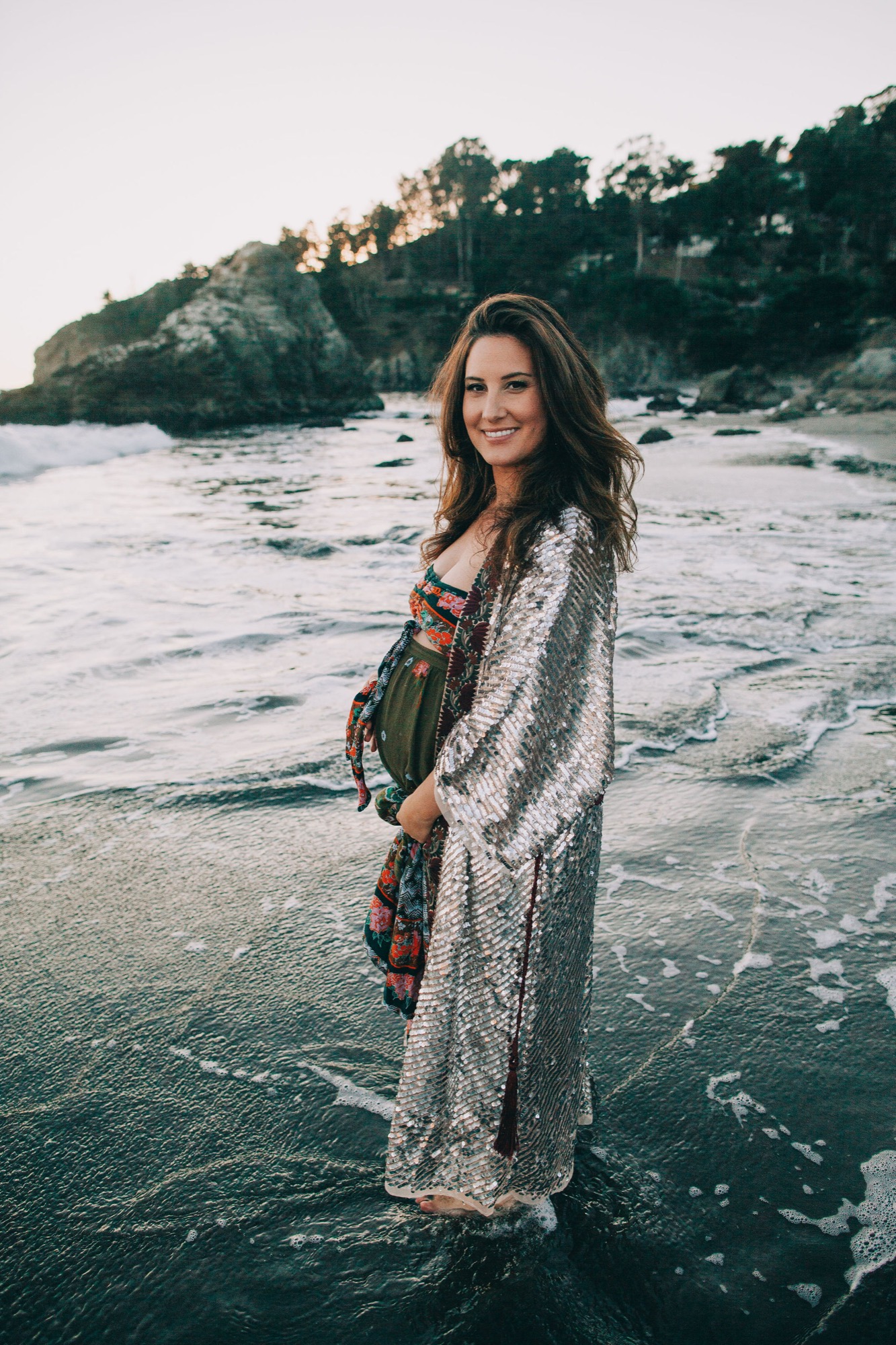 Pregnant woman in shimmering cardigan wades through shallow waves at sunset, the coastline glowing golden behind her