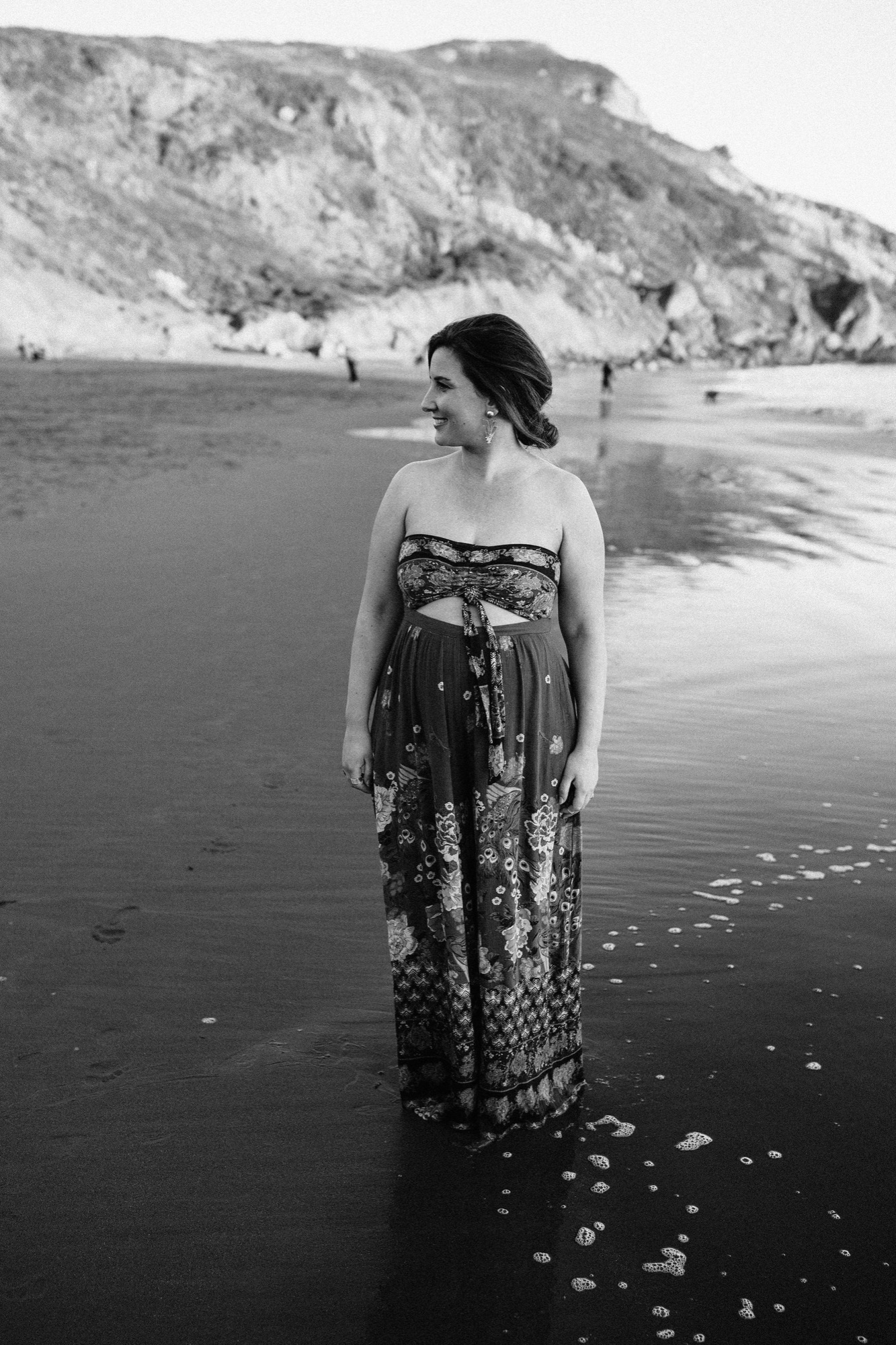 Woman in patterned wrap dress stands in shallow water at Muir Beach, gazing toward the horizon as waves move around her