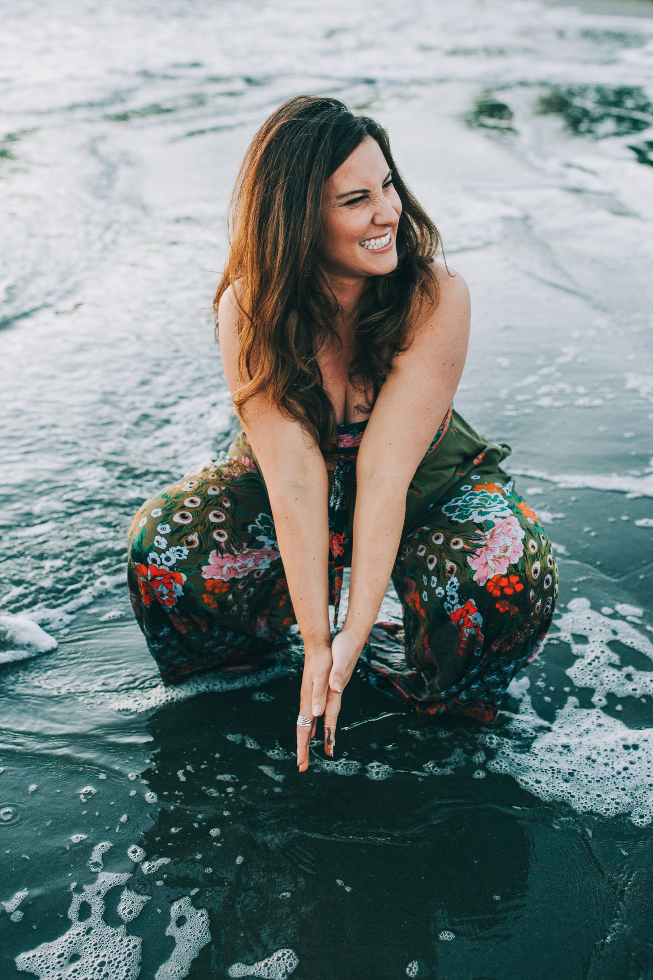 Pregnant woman in floral pants crouches in shallow surf, beaming with golden hour light catching her dark hair and bare shoulders