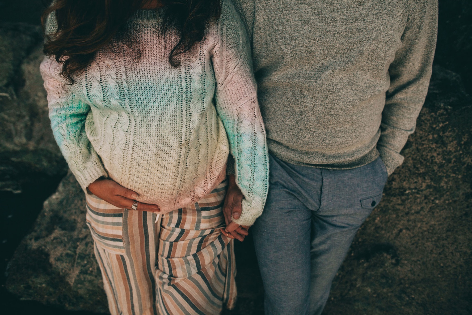 Expectant parents stand close together on rocky ground, her hand cradling her belly in a cream cable-knit sweater, his arm protectively around her