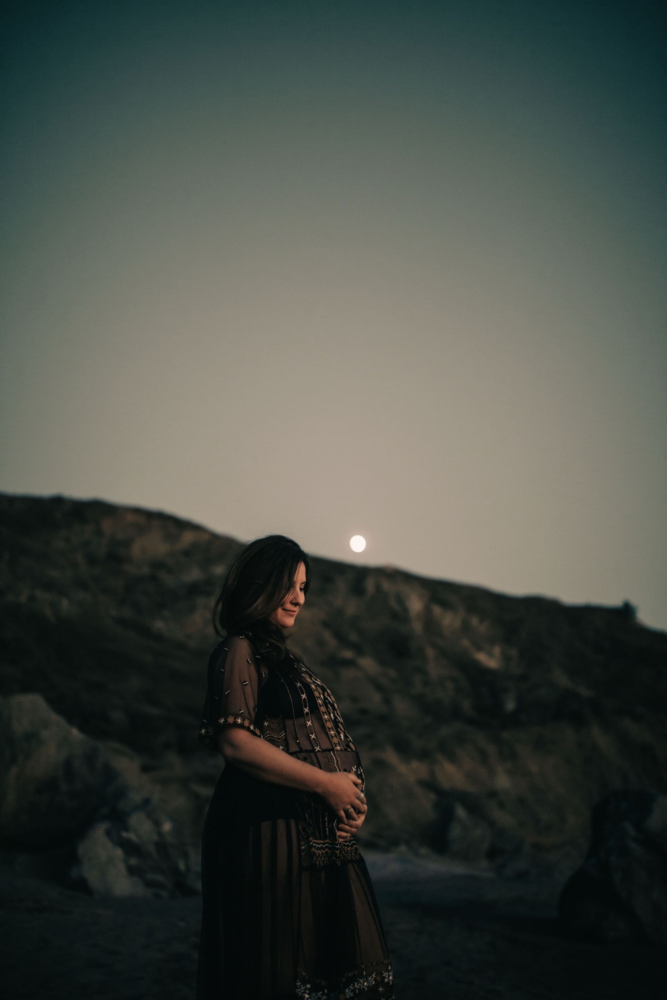 Pregnant woman in ornate black dress gazes over her shoulder as the full moon rises behind rocky hills at dusk