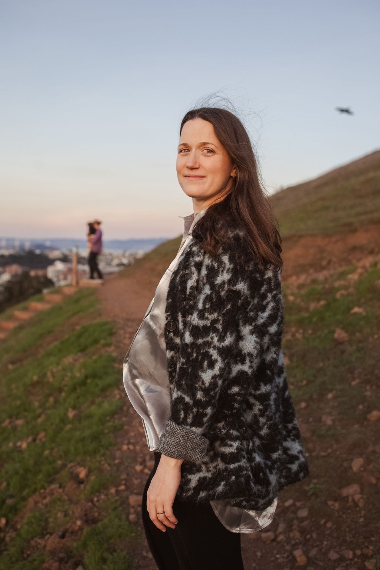 Pregnant woman in a black and white patterned wrap smiles over her shoulder on a windswept hillside at golden hour