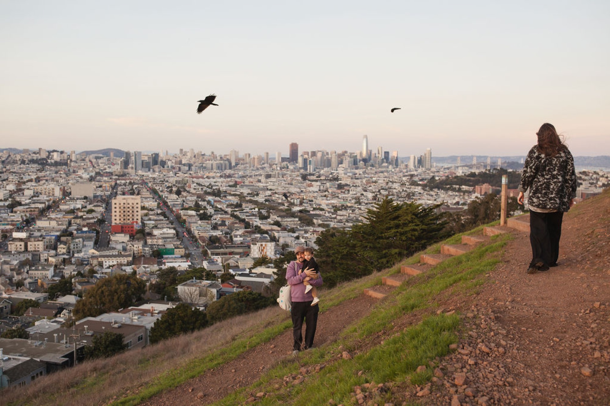 Pregnant woman stands on a grassy hilltop overlooking San Francisco's skyline at golden hour, while a couple embraces in the middle distance