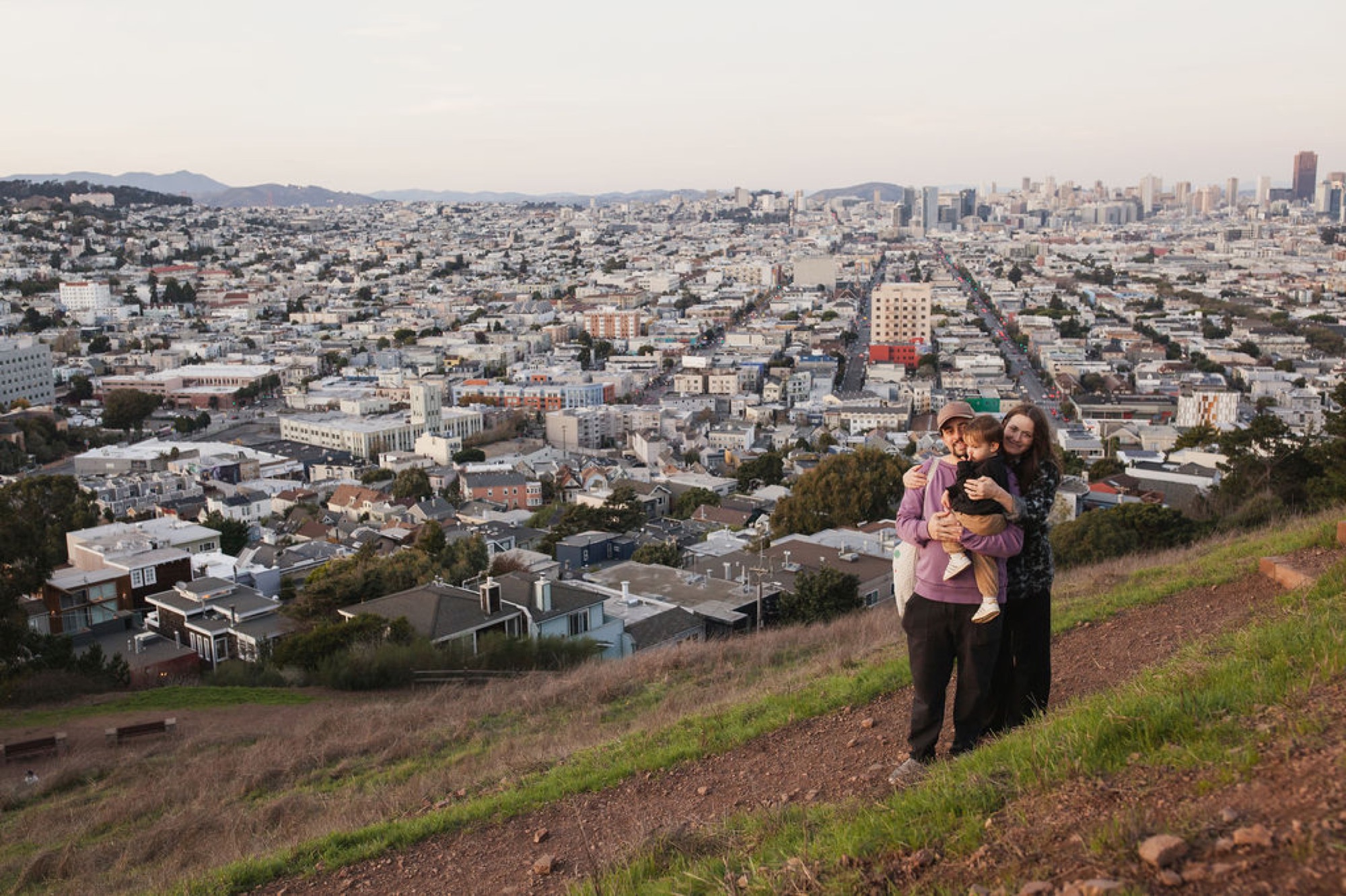 Expecting parents embrace on a windswept hillside overlooking San Francisco's sprawling cityscape, golden afternoon light illuminating the landscape