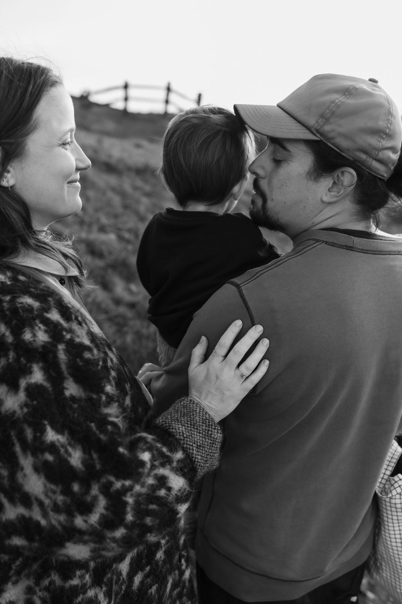 A couple holds their young child between them on a hillside, gazing down at their toddler in a moment of quiet connection