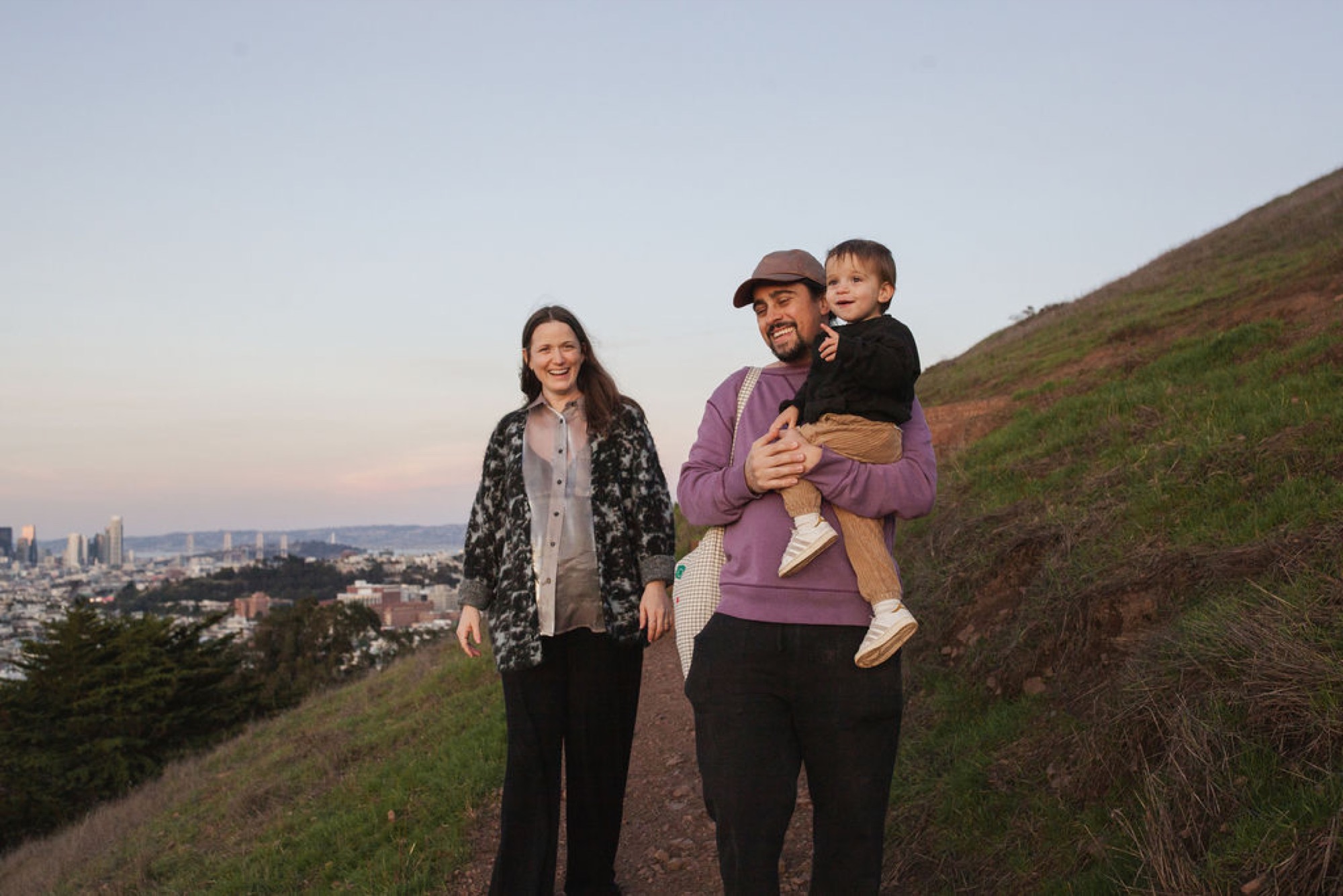 Family of three stands on a grassy hilltop at golden hour, with the man holding a young child while the woman walks beside them, city skyline visible in the distant background