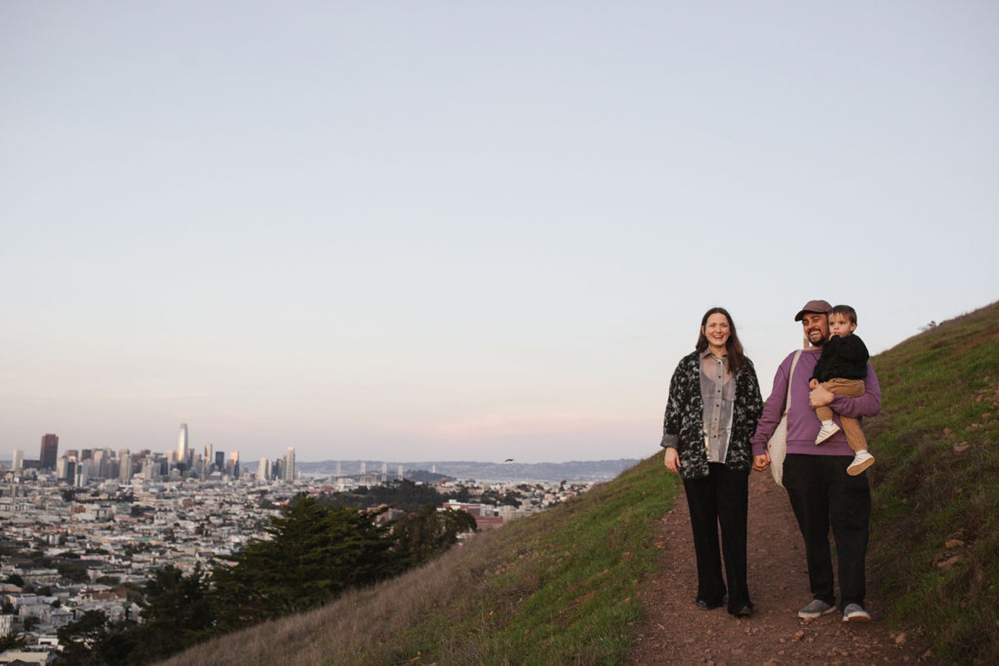 Two women walk side by side on a hillside trail with San Francisco's skyline glowing softly in the distance behind them