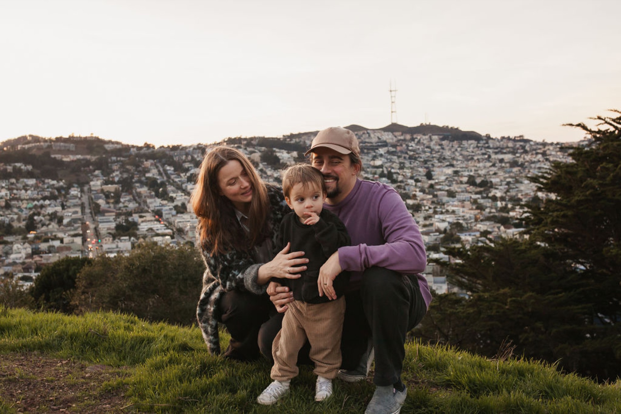 A family of three crouches together on a grassy hilltop overlooking San Francisco's dense hillside neighborhoods, their bodies angled toward each other in an intimate embrace