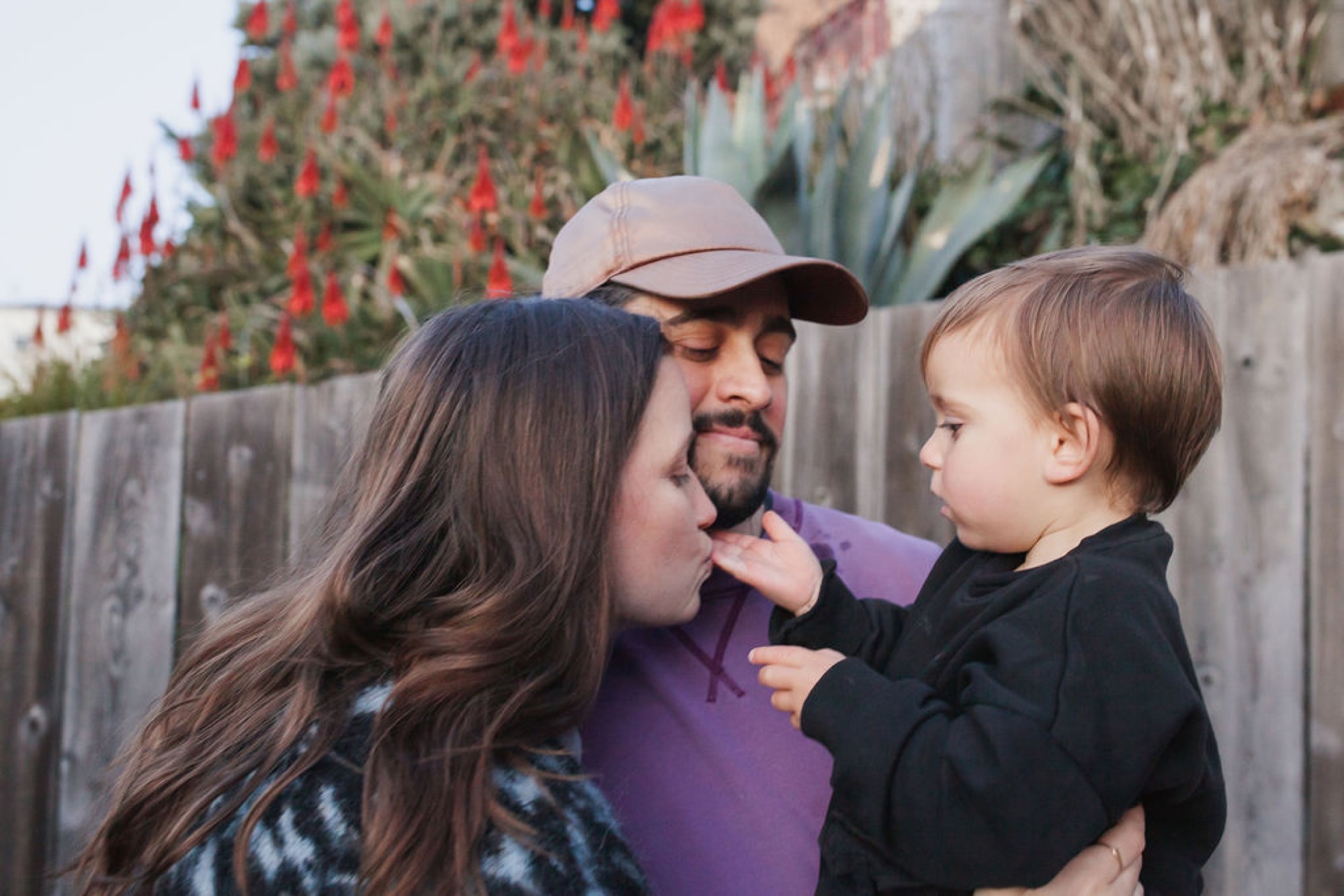 A man in a purple shirt and cap holds a toddler while a woman with long dark hair leans in close, their faces nearly touching in an intimate moment