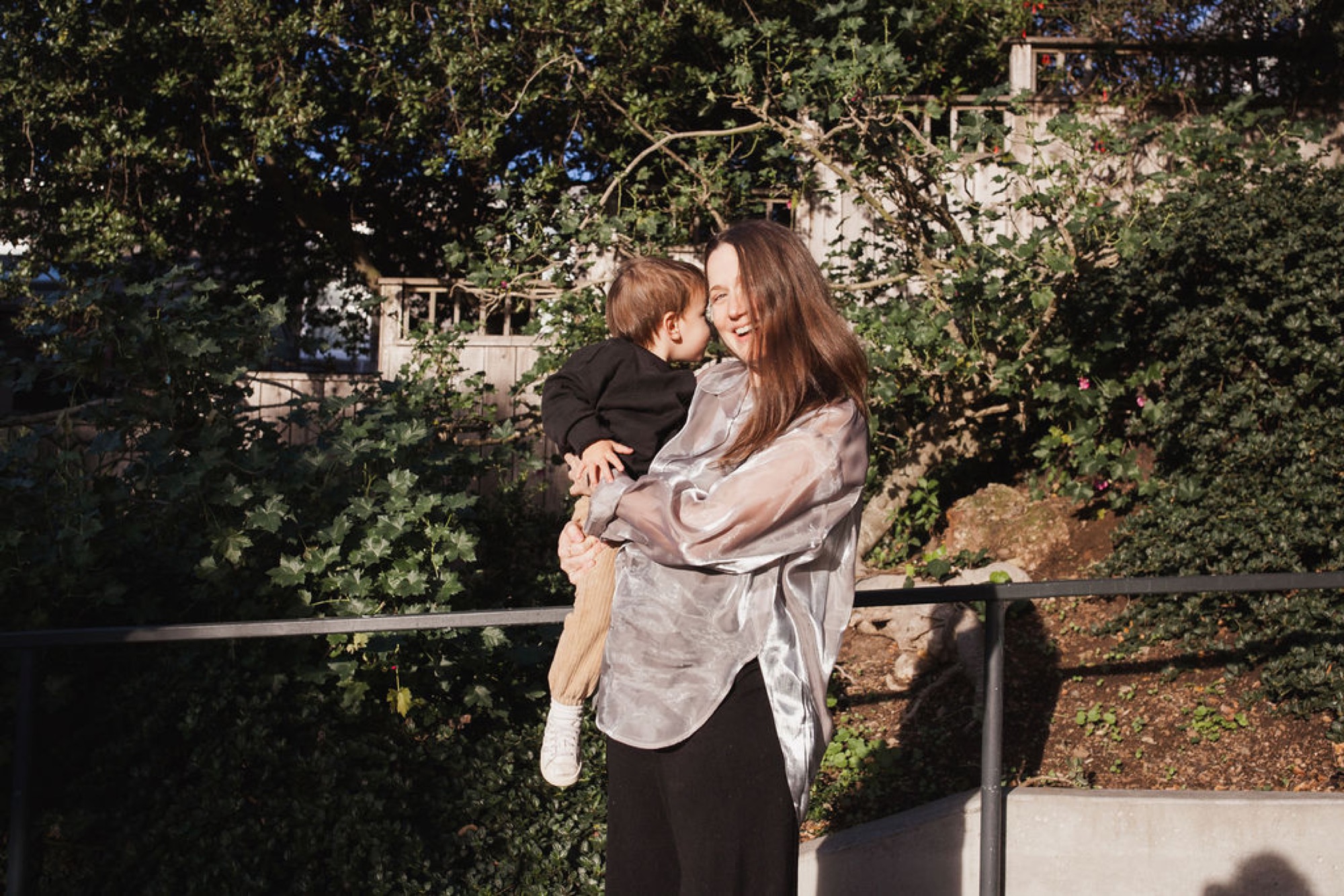 A woman in a light gray shirt holds her toddler son on a wooden railing, both smiling warmly with lush green foliage and ivy-covered structures behind them