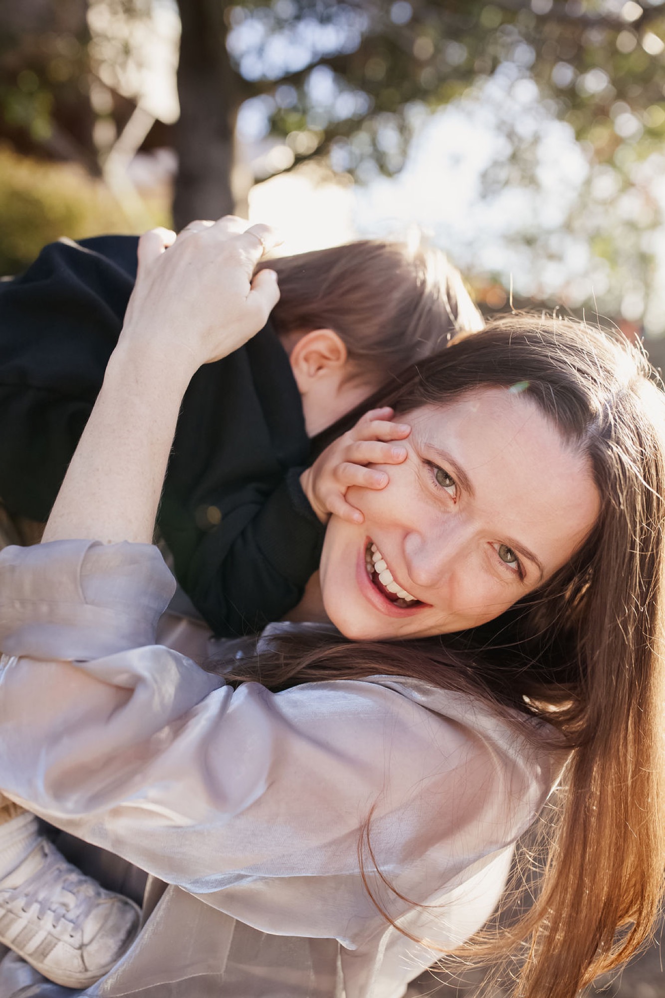 Woman in white laughs joyfully while man in dark jacket embraces her from behind in dappled sunlight