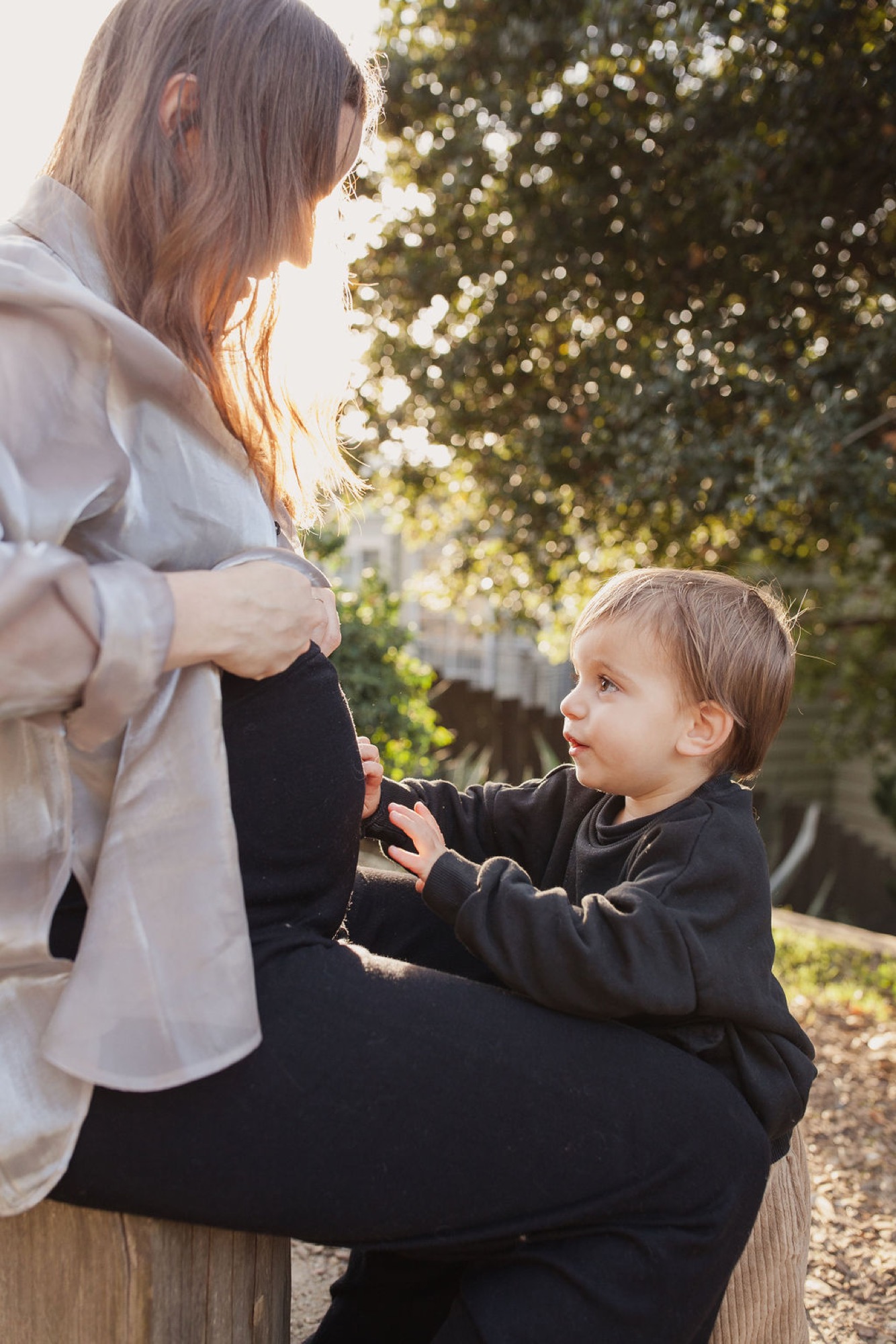 A toddler in dark clothing reaches toward a woman's pregnant belly in dappled sunlight beneath leafy green trees