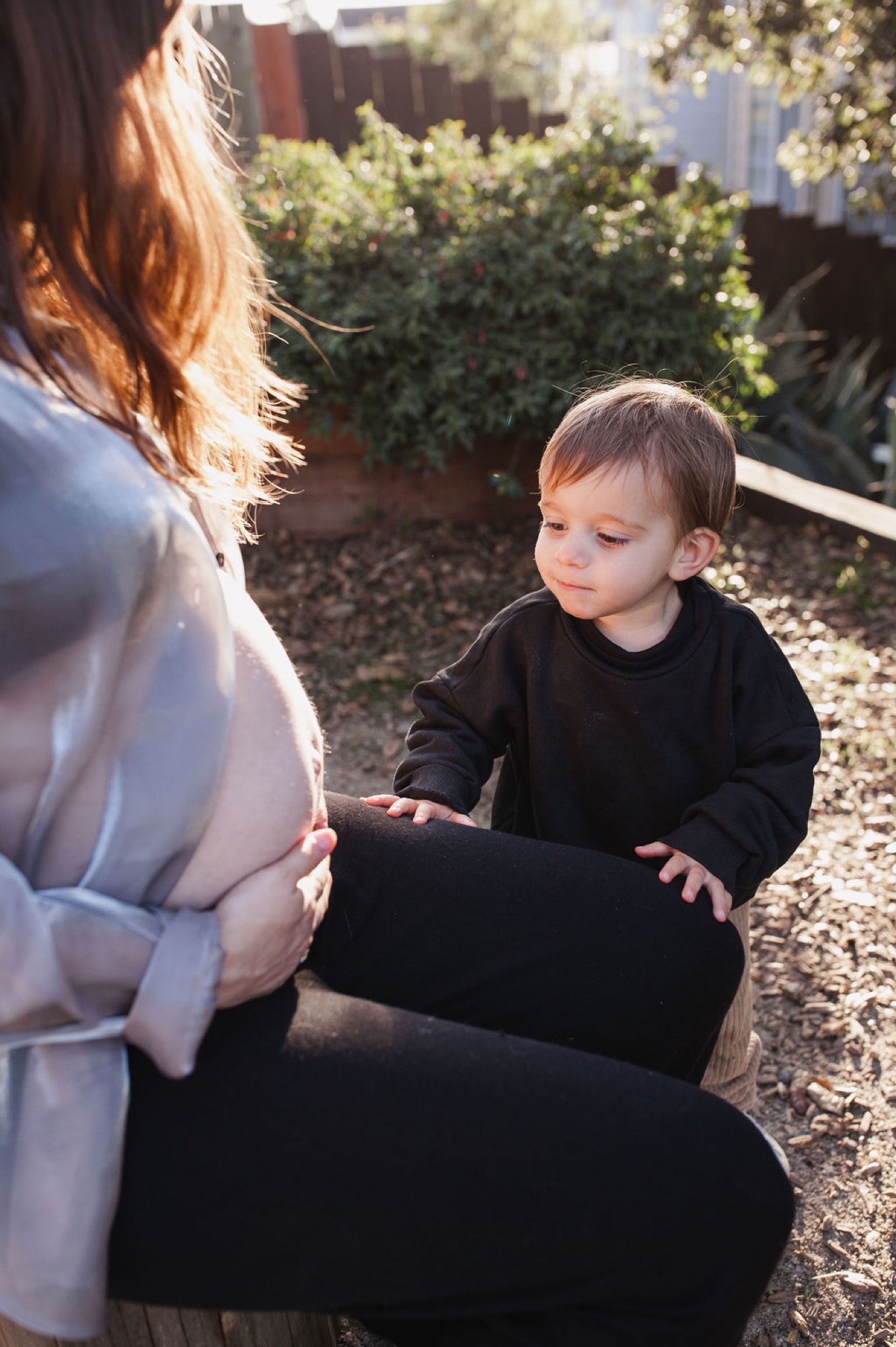 A toddler in black reaches toward his mother's pregnant belly in golden afternoon light, her reddish hair catching the sun in a backyard garden setting