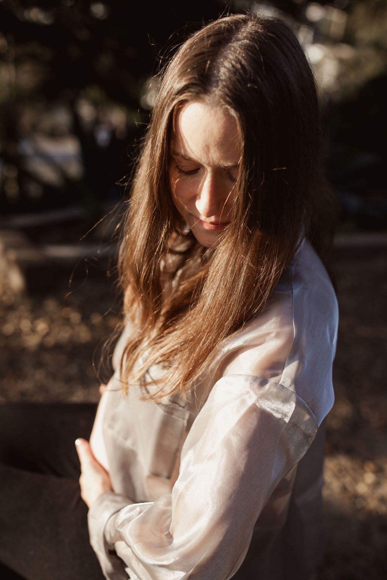 A woman in a crisp white shirt stands in dappled sunlight, gazing downward with a serene, contemplative expression as golden light filters through trees