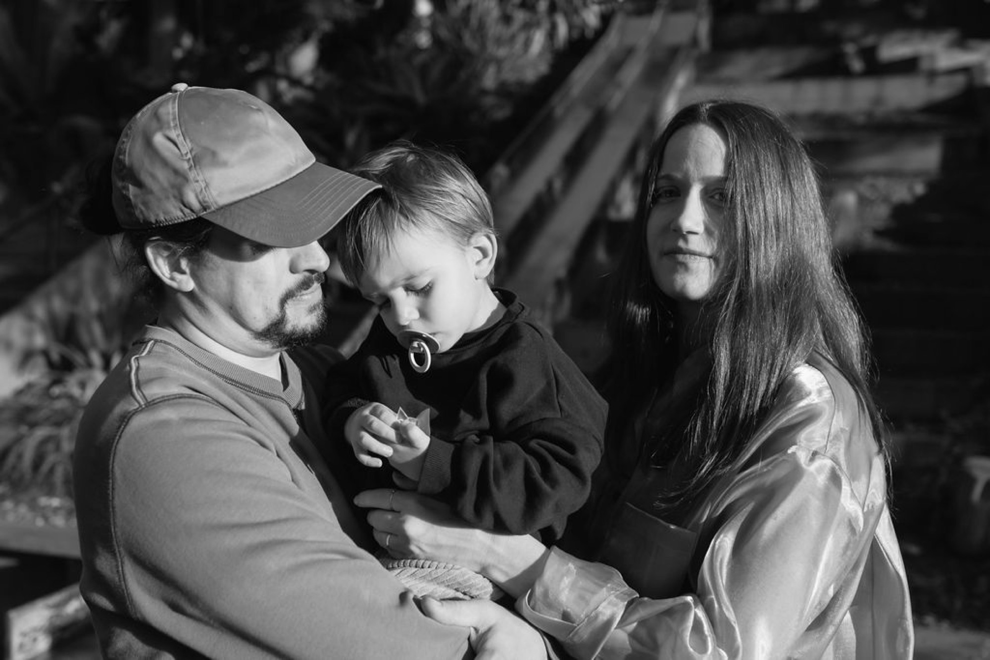 A man in a baseball cap holds his young child while a woman in long dark hair stands beside them in black and white