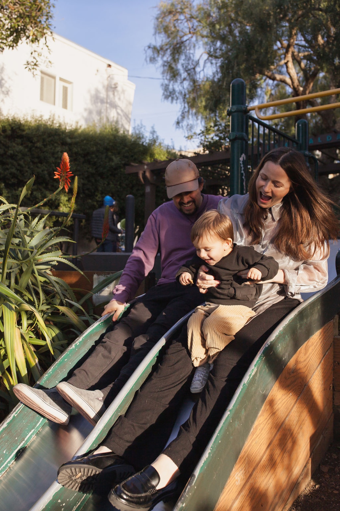 A beaming father and mother guide their toddler down a sun-dappled slide while tree shadows dance across the playground structure