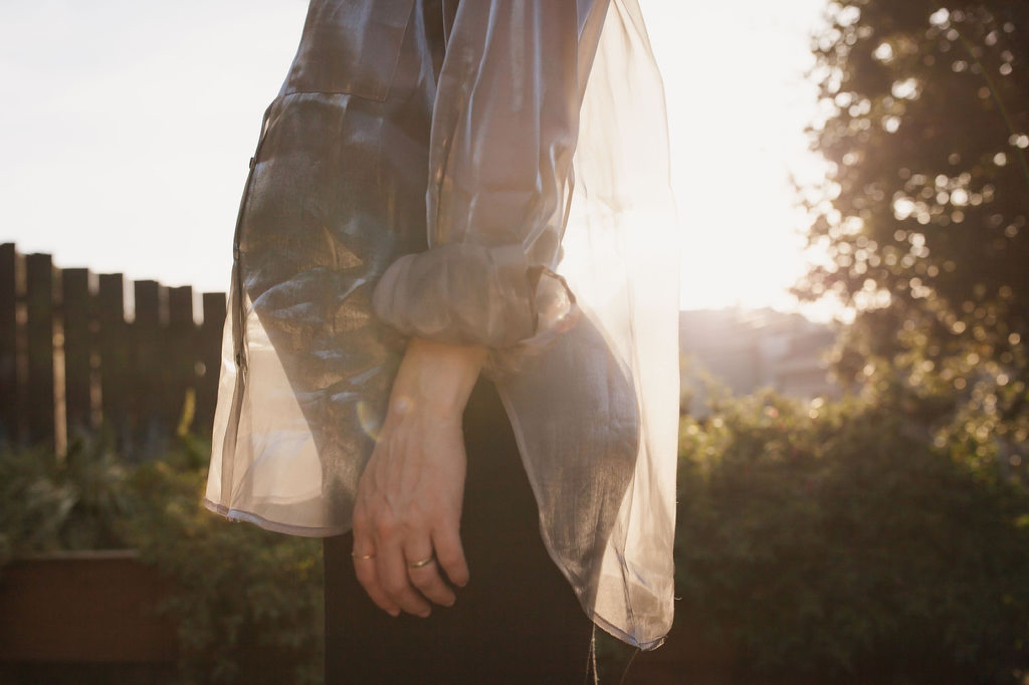 Pregnant woman in sheer overlay holds her draped belly against golden afternoon light filtering through foliage