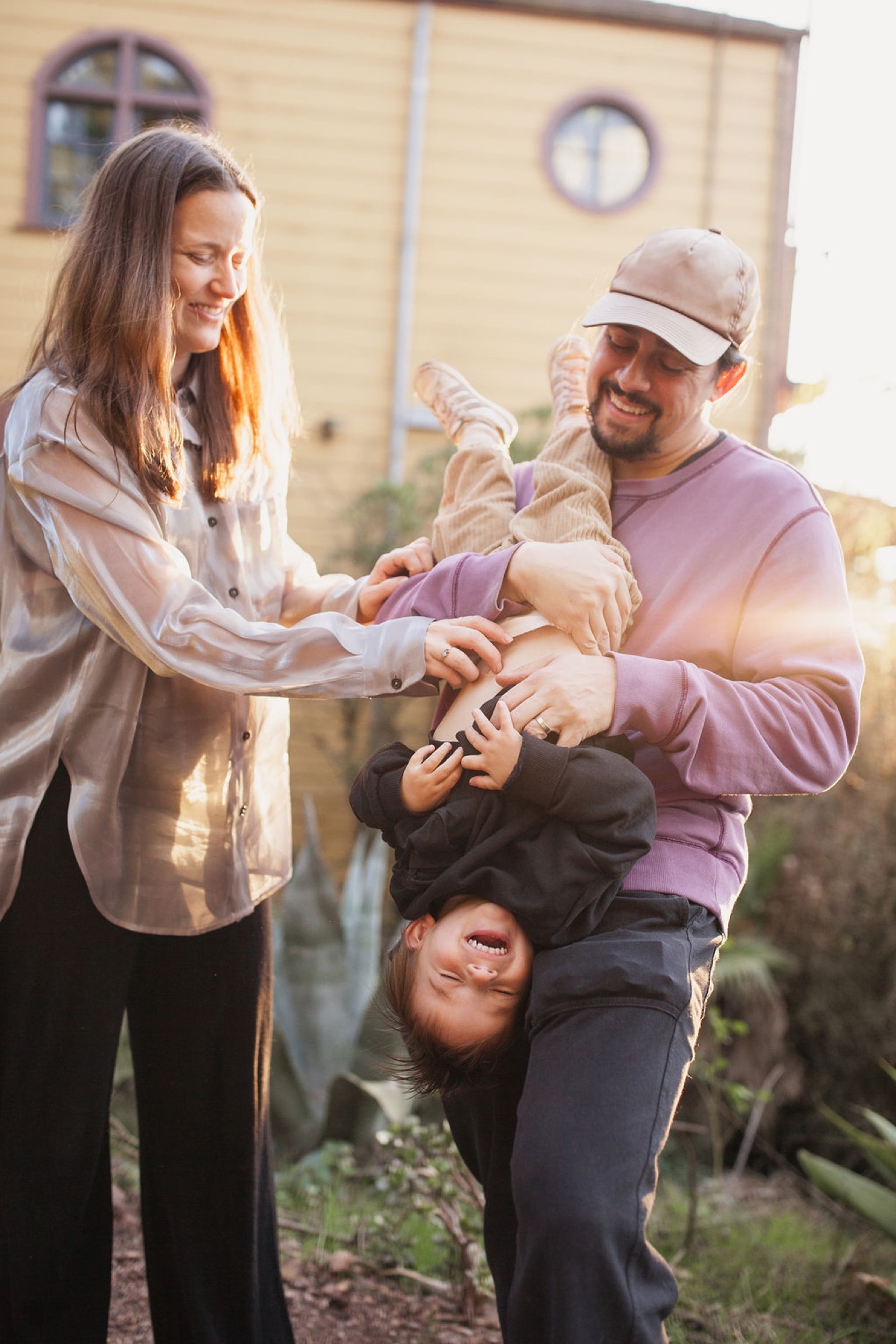 A man in a purple shirt and cap playfully holds his young child upside-down while a woman in a tan jacket smiles beside them in golden afternoon light