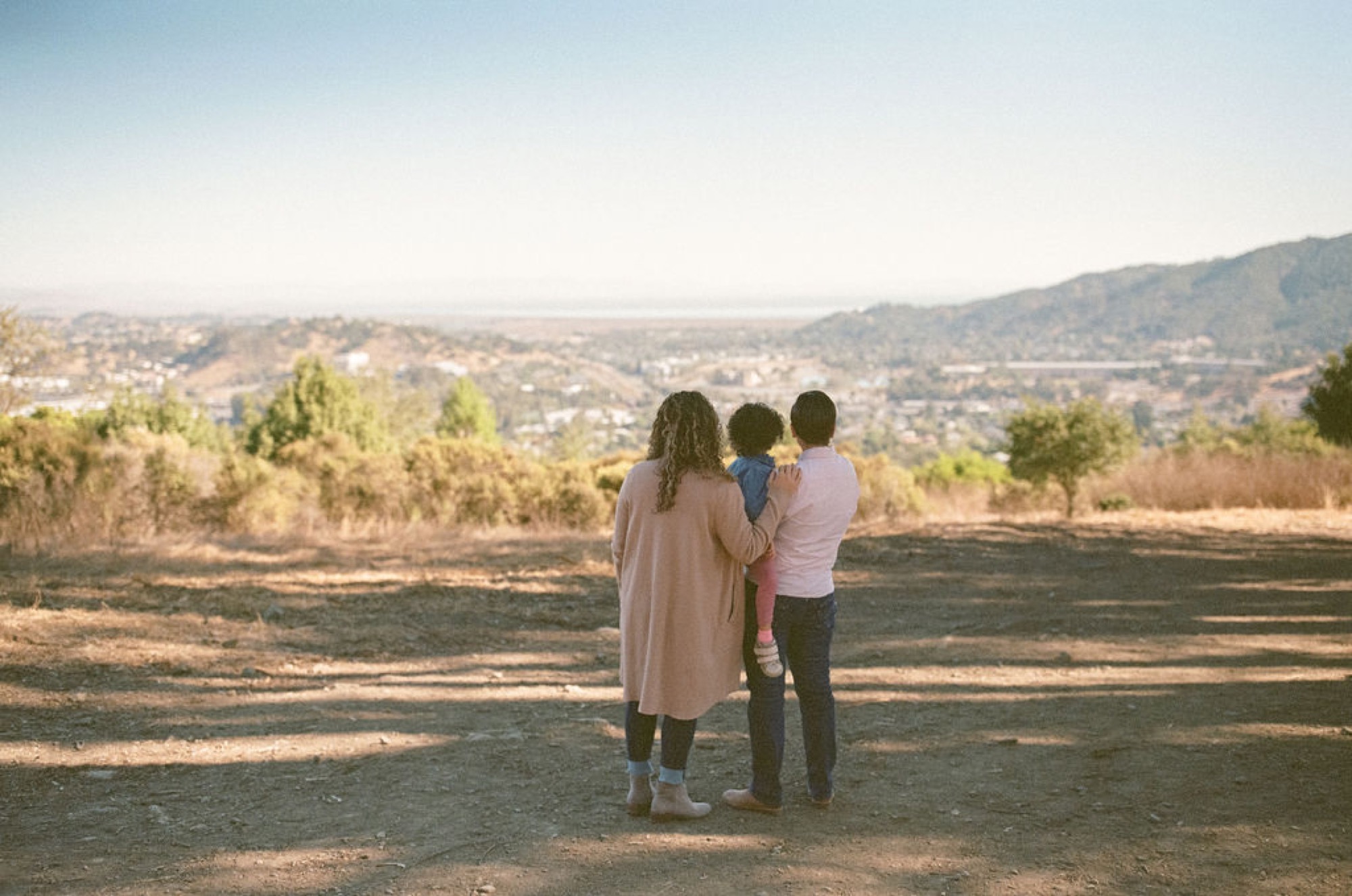 Two women hold their young child between them, gazing across a sprawling valley dotted with homes and distant hills under soft afternoon light