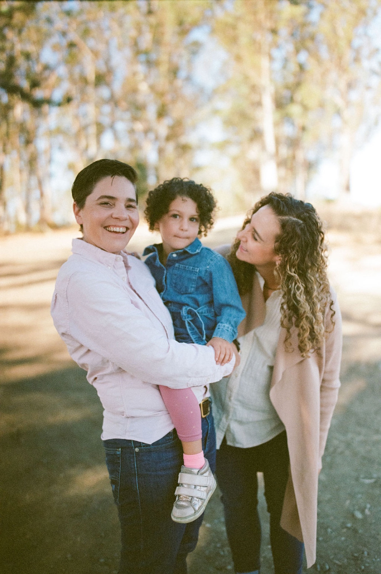 Two adults and a young child stand together on a tree-lined path, the person on the left beaming at the camera while holding the child in denim