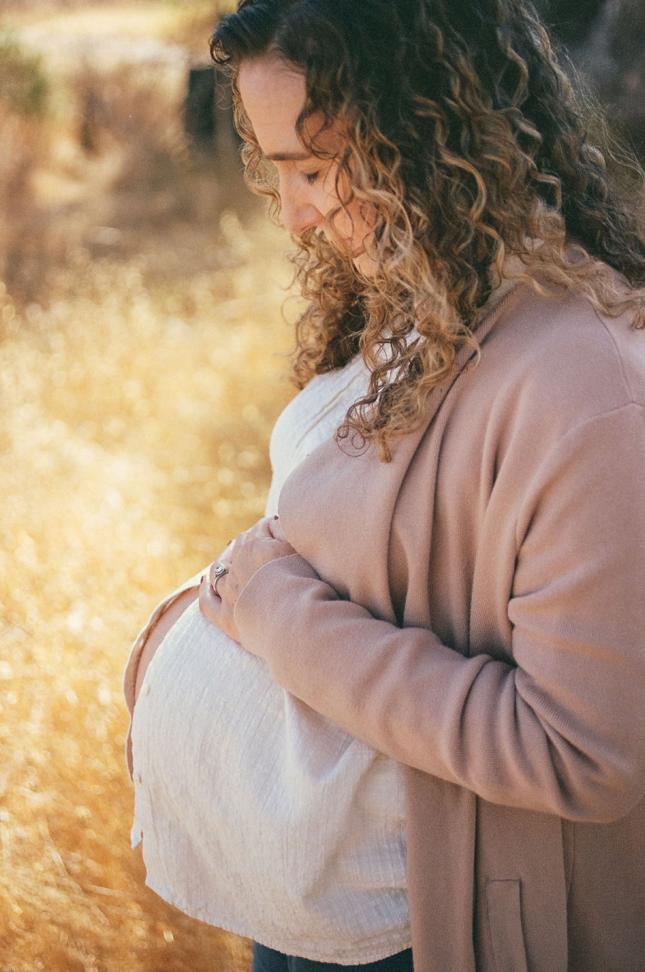Pregnant woman in profile gazes downward, cradling her belly with both hands, warm golden light illuminating her curled hair and peaceful expression