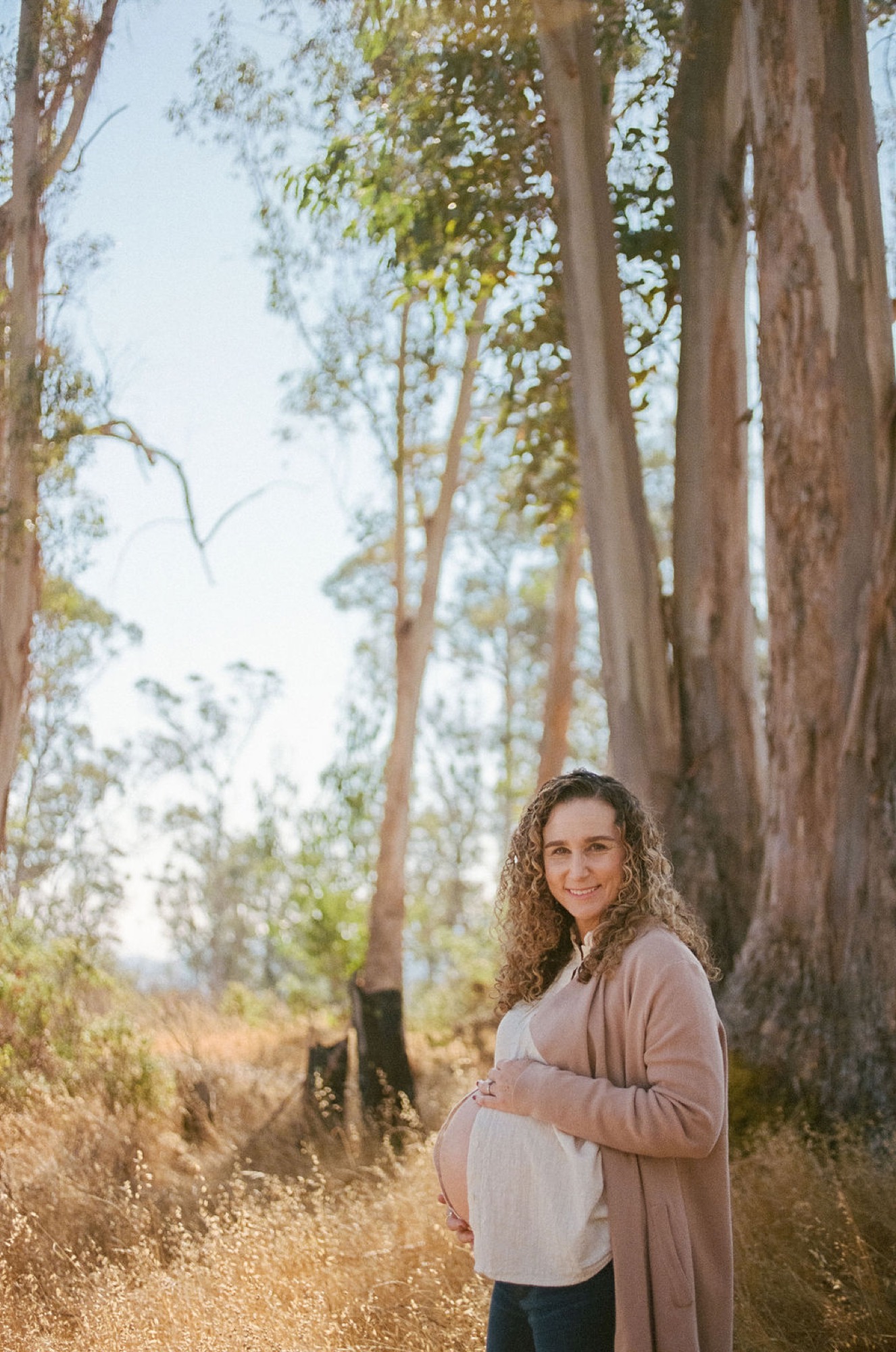 Pregnant woman with curly hair smiles warmly while cradling her belly beneath towering eucalyptus trees, golden afternoon light filtering through the canopy