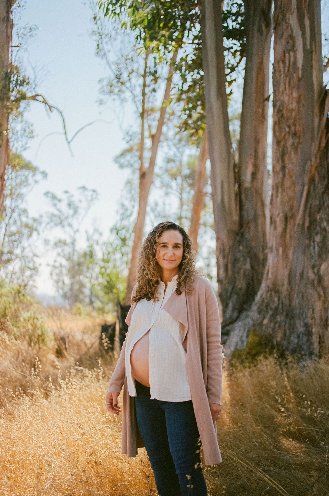 Pregnant woman with curly hair stands centered on a sunlit dirt trail between towering eucalyptus trees, wearing a cream top and taupe cardigan, gazing softly toward the camera